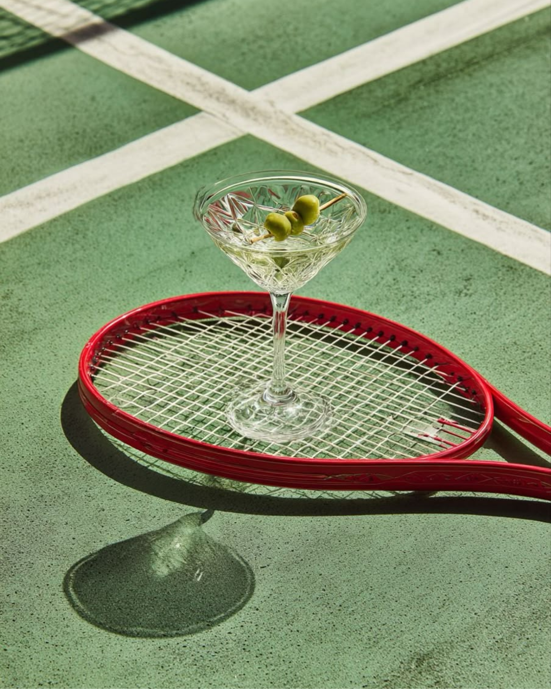 A martini glass with green olives on a toothpick, placed on a red tennis racket, casting a shadow on a green tennis court.