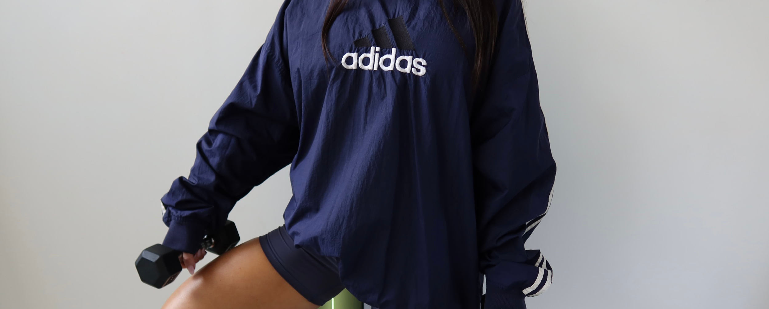 Person wearing an Adidas navy sports jacket holding a black dumbbell, with a plain light-colored background.