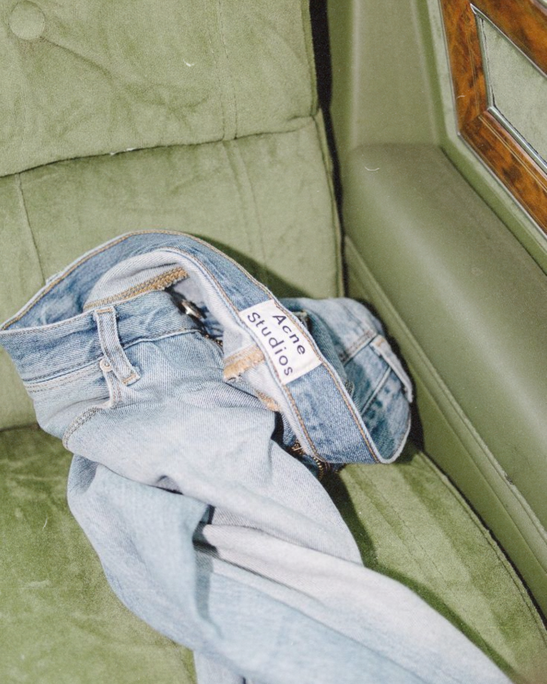 Close-up of denim jeans with a clothing label, lying on a green surface near a wooden and metal framed piece of furniture.