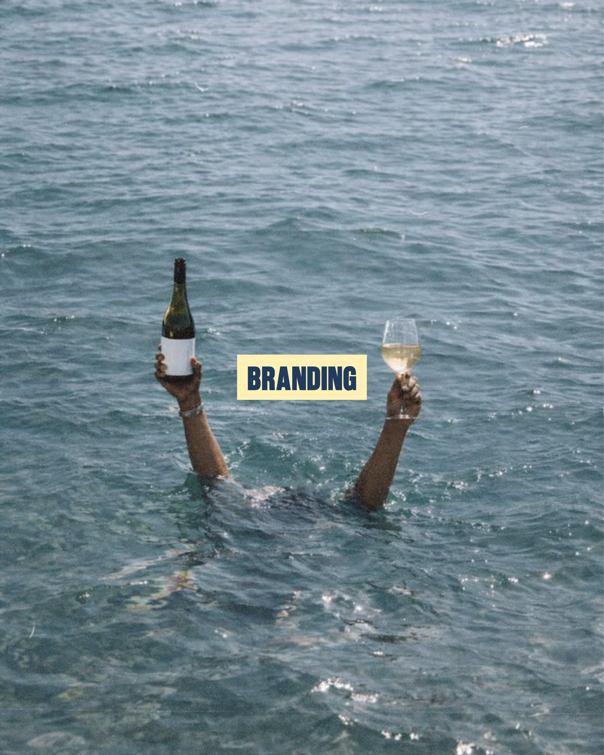 Two hands emerge from the water holding a bottle of wine and a glass of wine against a backdrop of a large body of water.