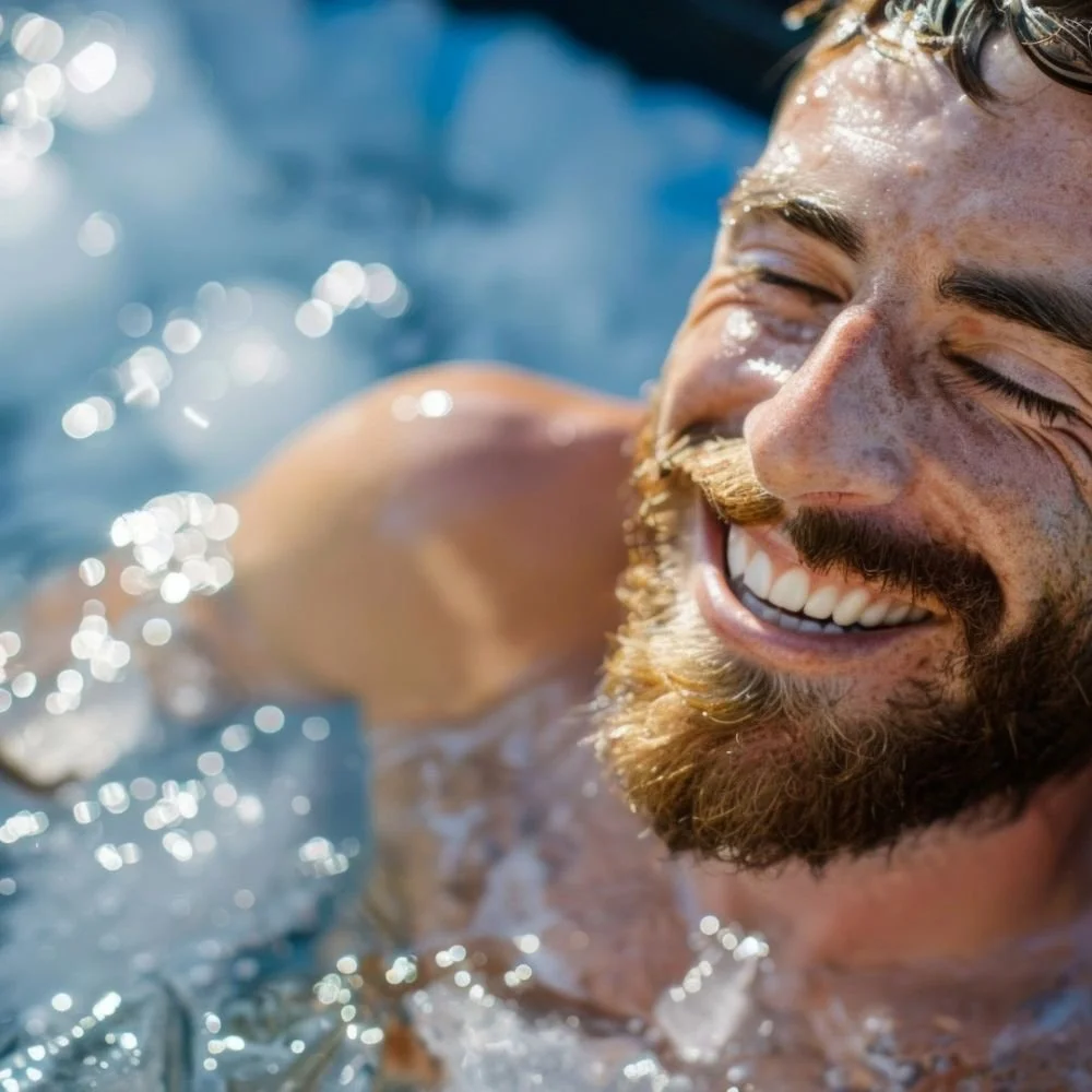 A man with eyes closed and laughing while sat in ice bath water, visible water droplets and sunlight reflections.