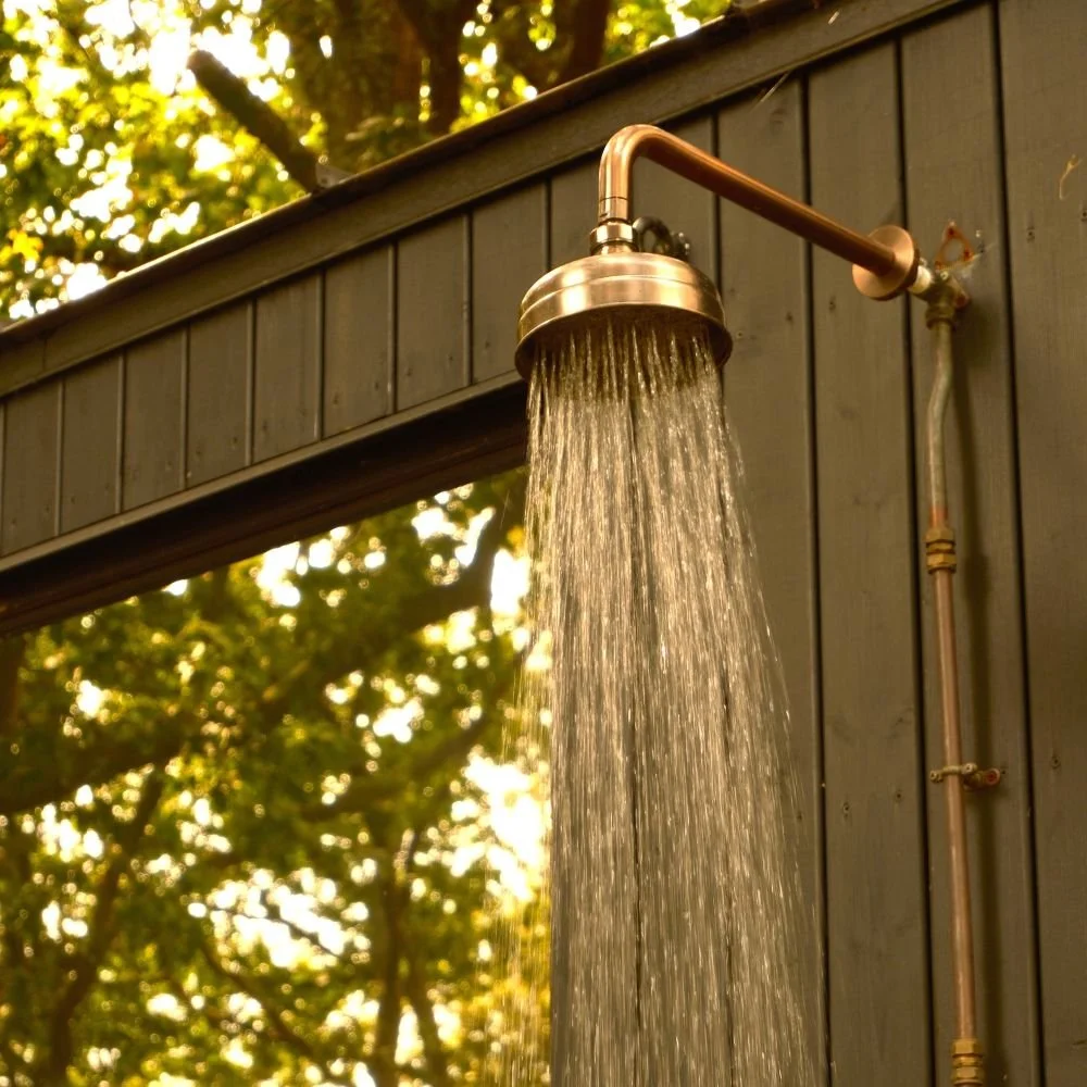 An outdoor shower with water running, mounted on a dark wooden sauna exterior wall with trees and sunlight in the background.