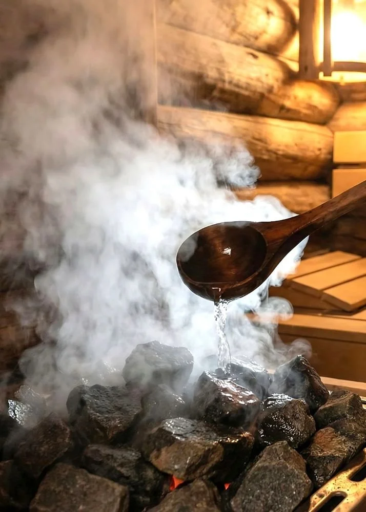 Hot stones with steam rising in a sauna, with a wooden ladle pouring water onto the stones, and wooden logs in the background.