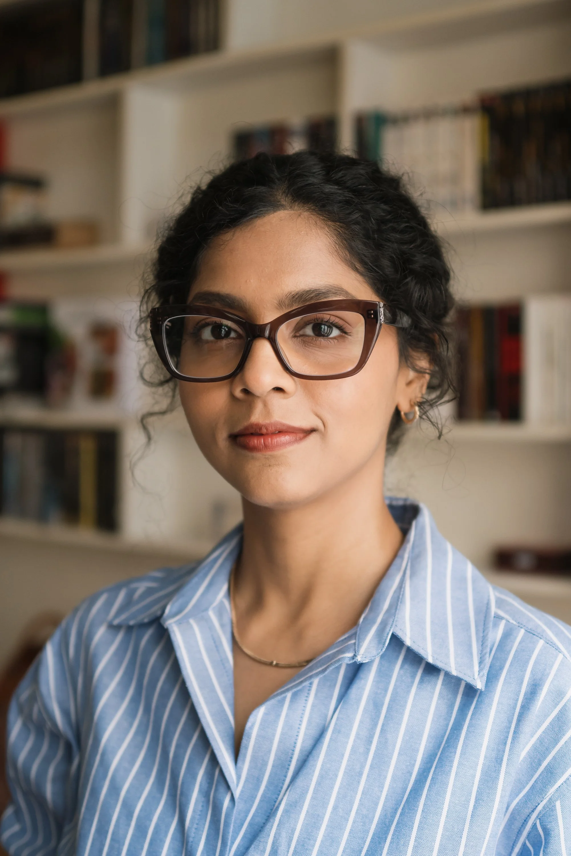 A young woman with dark curly hair, wearing glasses and a blue striped shirt, standing in front of a bookshelf.