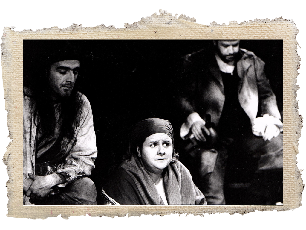 Sara Ford Black and white photo of three people, one woman with a worried expression, two men, one on the left with long hair and a hat, and another on the right with dark hair and a beard, sitting around.