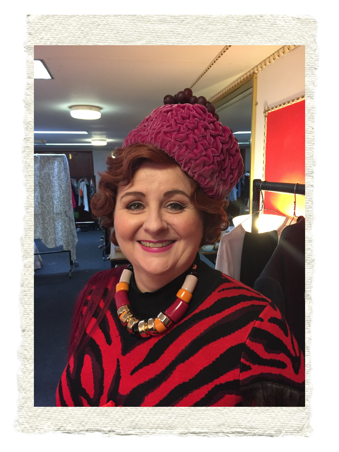 Sara Ford Woman with red curly hair smiling, wearing a red and black patterned dress, a bold necklace with large beads, and a large pink textured hat with grapes on top, in a room with clothing racks and warm lighting.