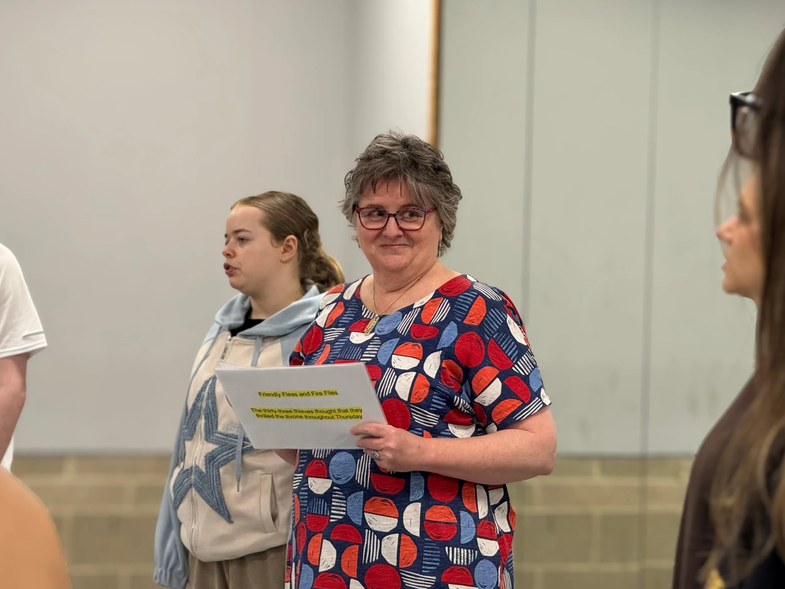 Sara Ford A woman with short gray hair and glasses holding a piece of paper and engaging in conversation at an indoor event, with other women visible in the background.