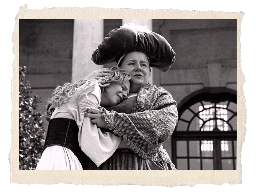 Sara Ford A woman and a young girl embracing, with the woman wearing a loose scarf on her head and a textured shawl, in front of a building with a large window and an arch.