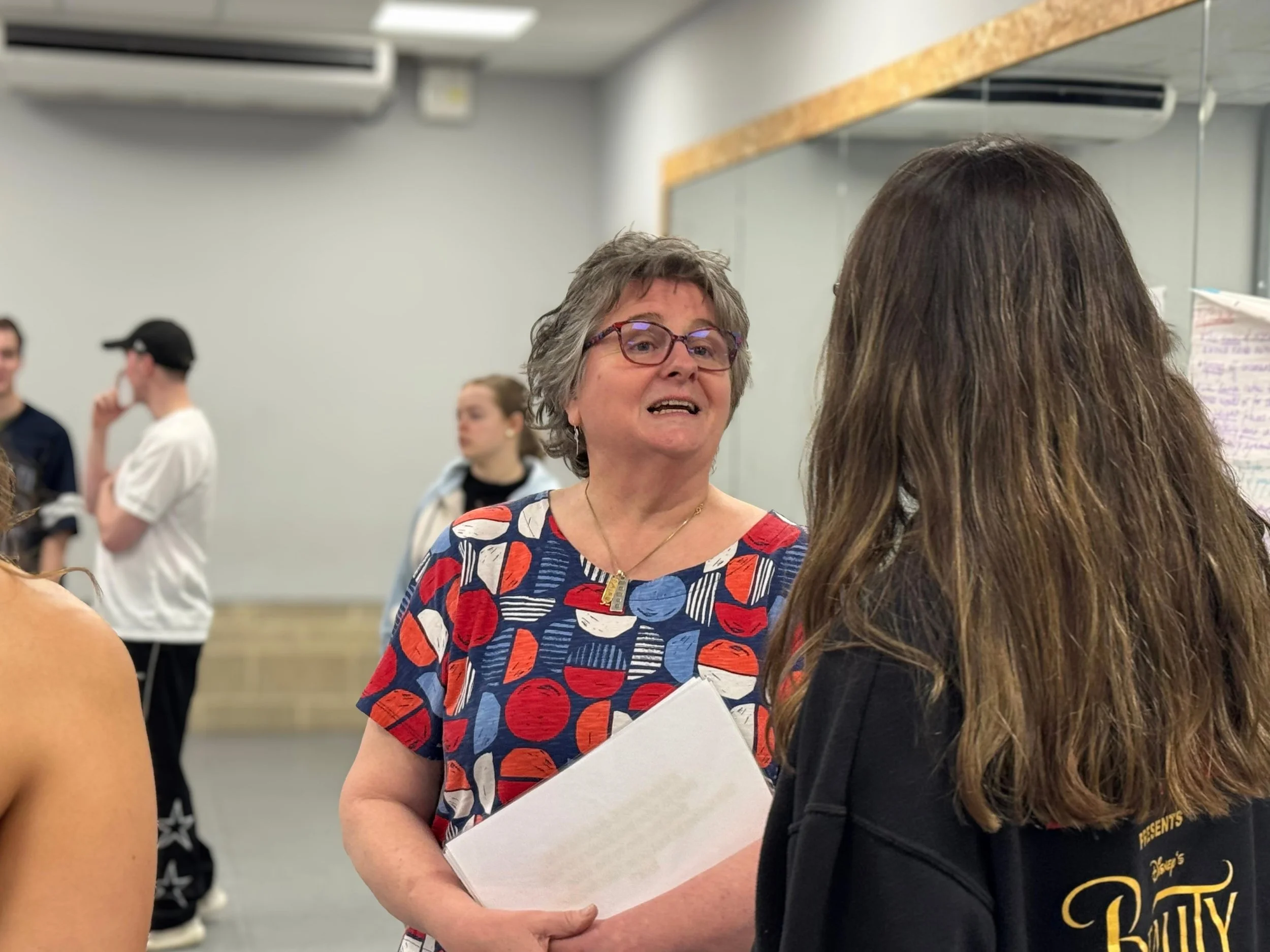 A woman with short gray hair and glasses is conversing with another woman who has long brown hair. The woman with gray hair is holding a white folder and appears to be speaking. The setting appears to be a classroom or conference room with several people in the background, some of whom are engaged in conversations.
