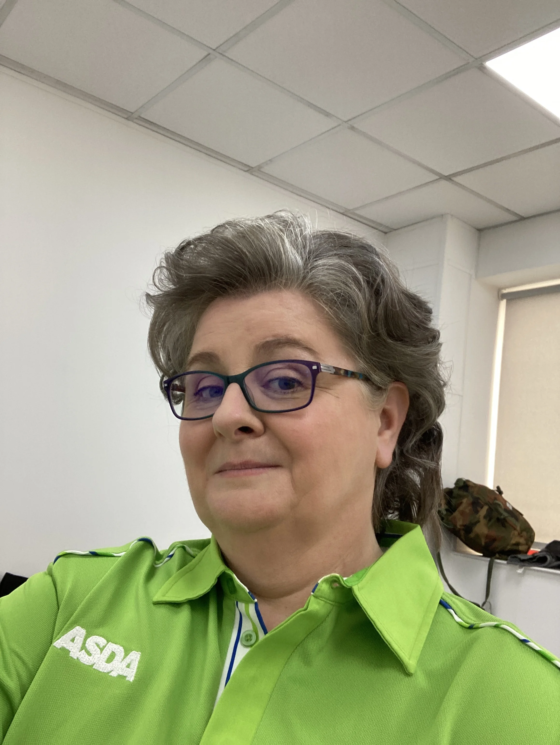 Sara Ford Close-up of a woman wearing glasses and a bright green uniform with the words 'ASQAA' visible. She has gray, styled hair and is indoors, with ceiling tiles and a window in the background.