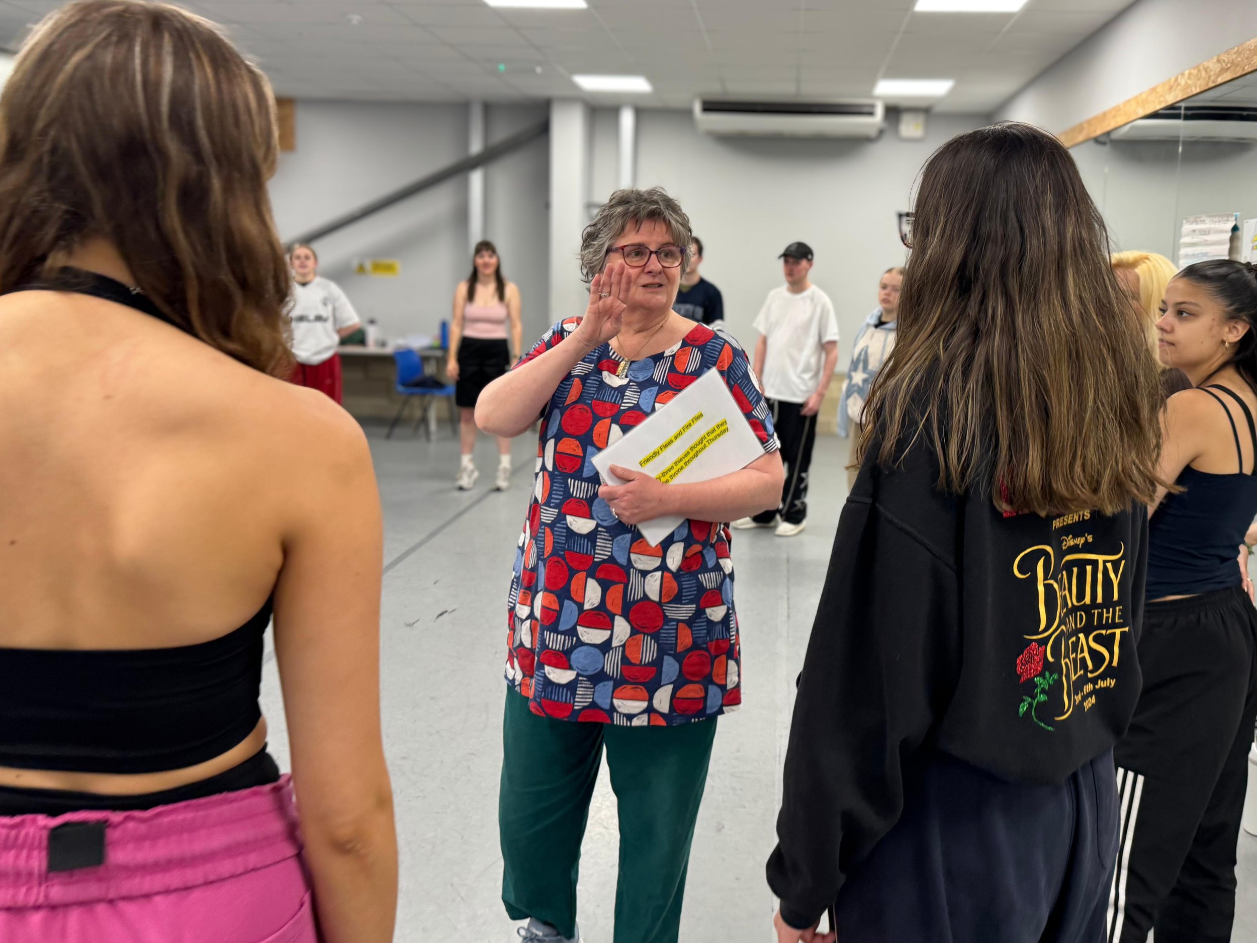 A woman with short gray hair and glasses gestures with her right hand while holding papers and speaking to a group of diverse young women in a gym or dance studio.