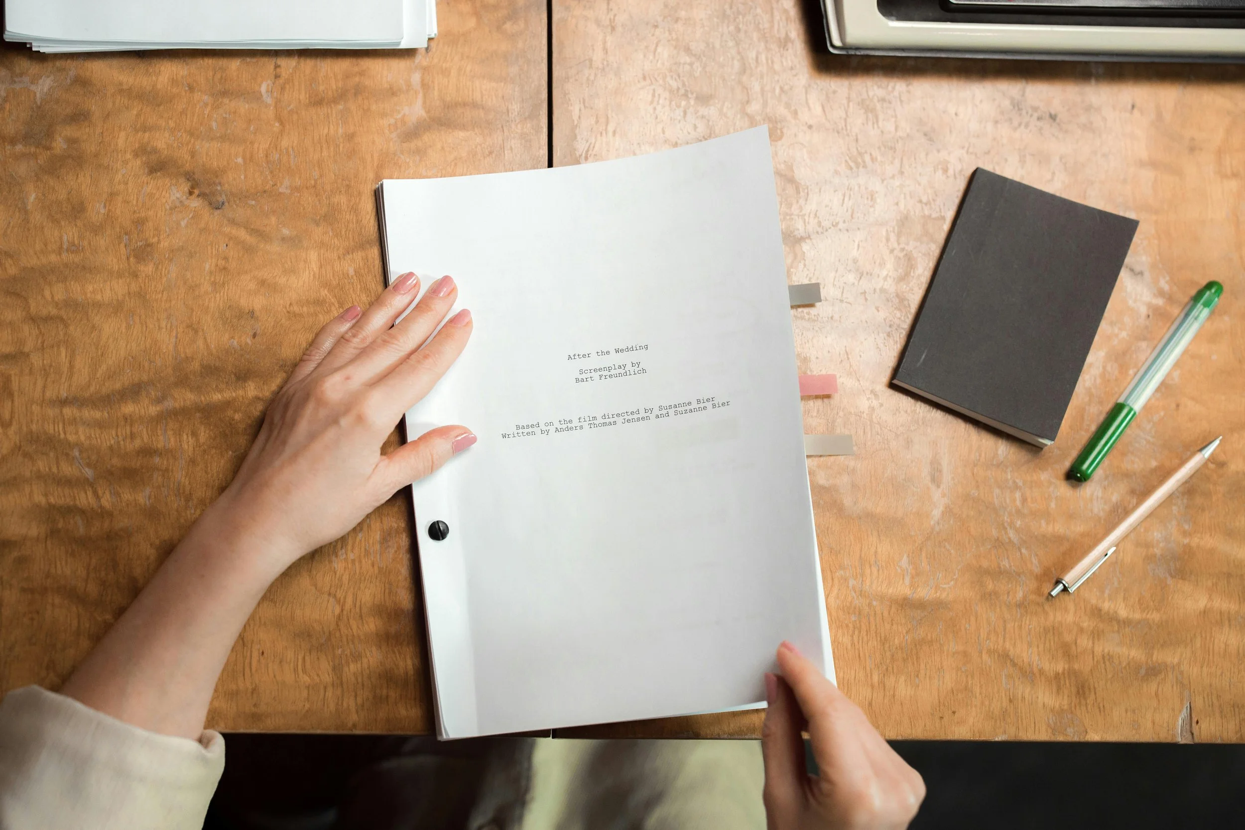 A person holding a script booklet titled 'After the Wedding' on a wooden table with office supplies around.