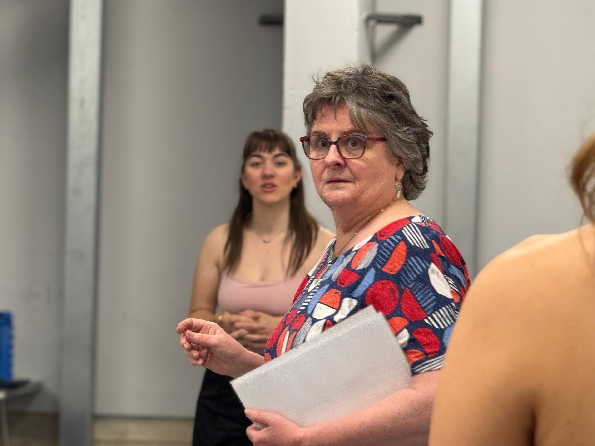 Sara Ford Women engaged in conversation in an indoor setting, one woman with short hair, glasses, and a colorful patterned shirt. Another woman with long brown hair and a light top is blurred in the background.