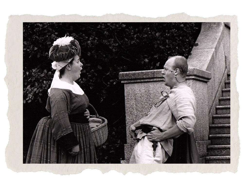 Sara Ford A black-and-white photo of a woman dressed in historical clothing, talking to a man wearing casual historical attire. The woman holds a basket, and the man has his hands on his hips, standing outside near a staircase and bushes.