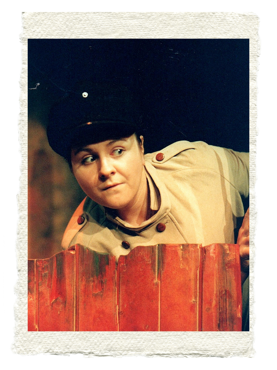 Sara Ford A woman dressed as a soldier in a beige uniform and black hat, peeking over a weathered red wooden fence.