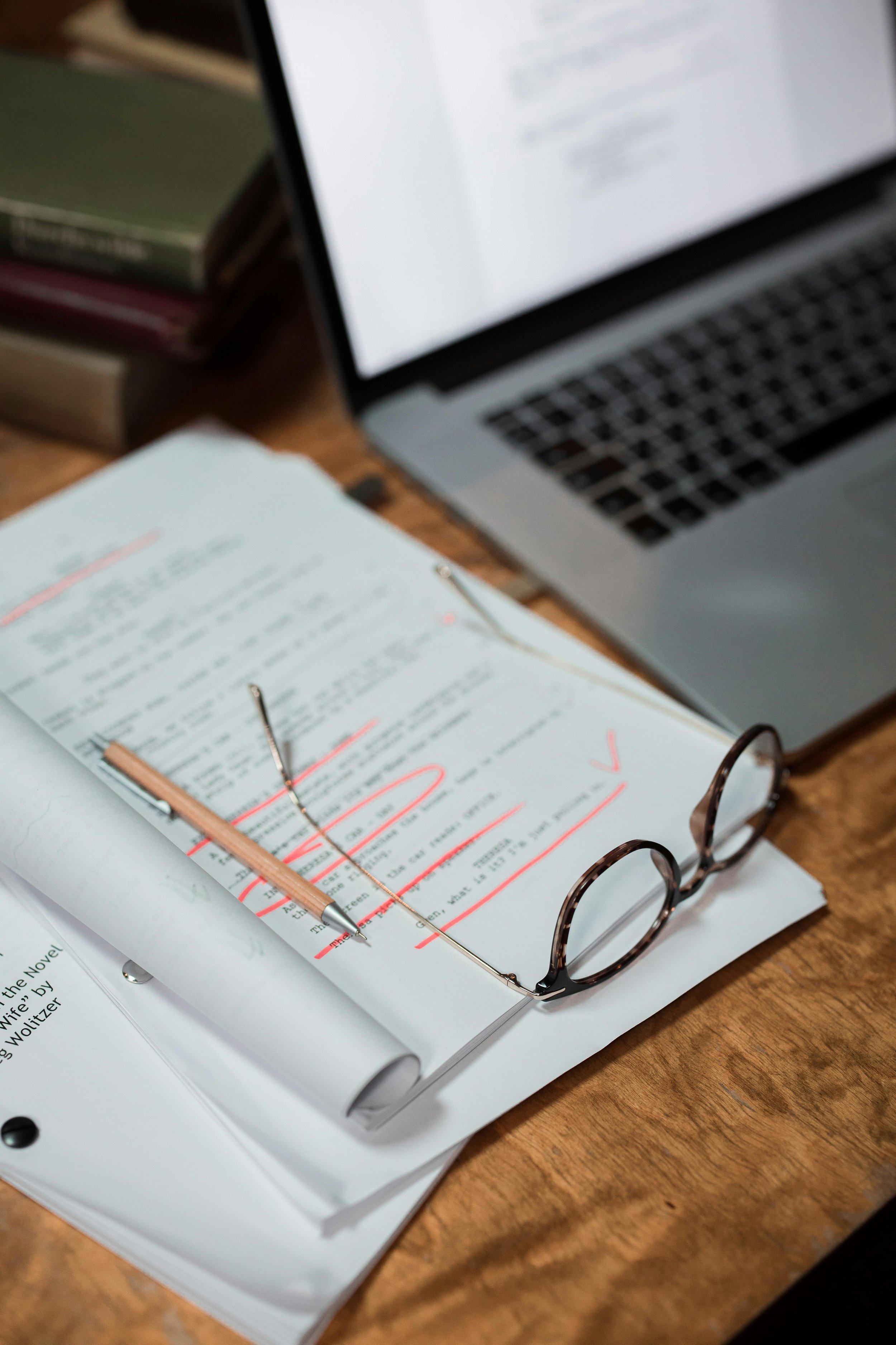 Open book with red highlighted text, a pair of eyeglasses, a wooden pencil with a silver tip, on a wooden desk, with a laptop and stacked books in the background.