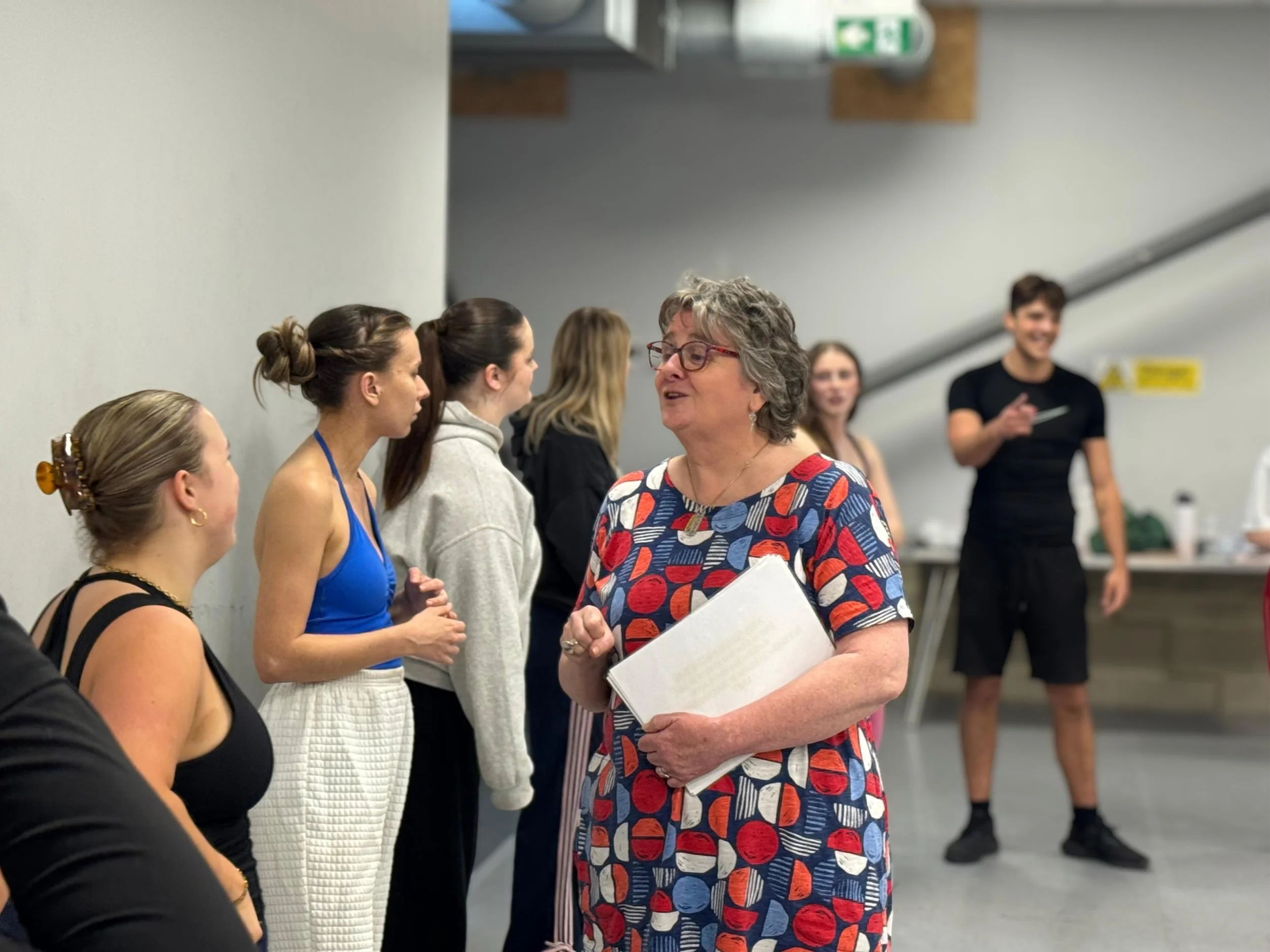 A group of women and a woman with gray hair and glasses are engaged in a conversation in an indoor setting. Other people are blurred in the background.