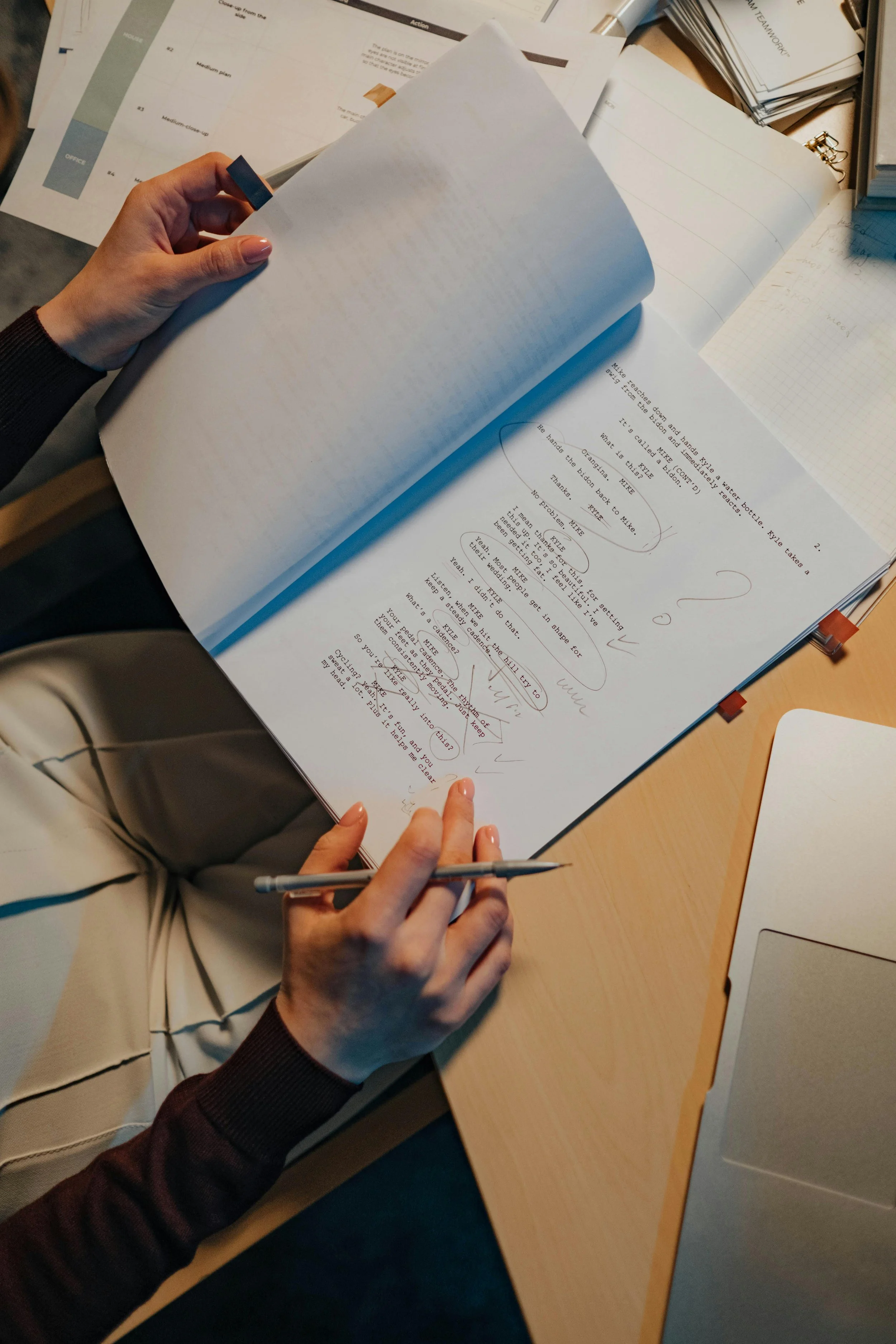A person with painted nails holding a pen and flipping through pages of a script or manuscript with handwritten notes and doodles on a desk filled with printed papers and notebooks.