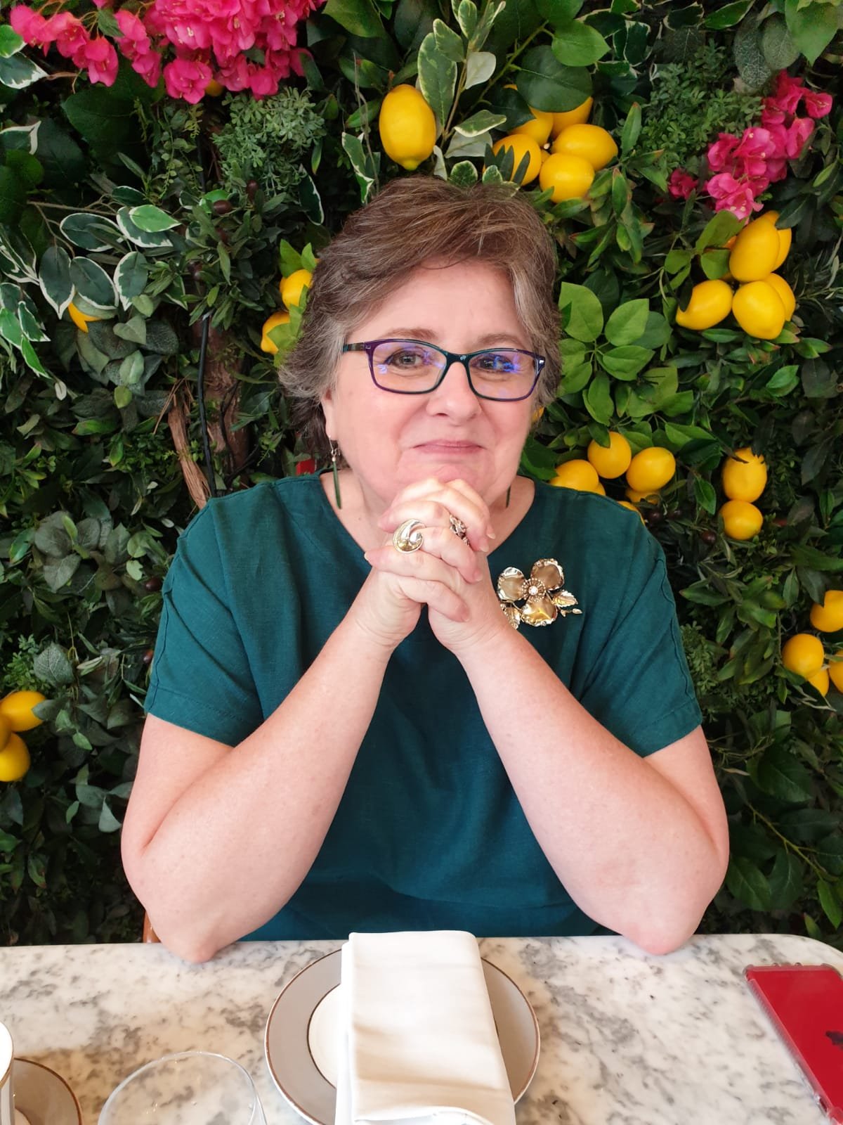 A woman with short brown hair, wearing glasses, a teal shirt, and a decorative brooch, sits at a marble table with a white napkin and glassware, clasping her hands together in front of her chin. She is in front of a lush green background filled with various plants and yellow lemons, with pink flowers, and appears to be in a garden or botanical setting.