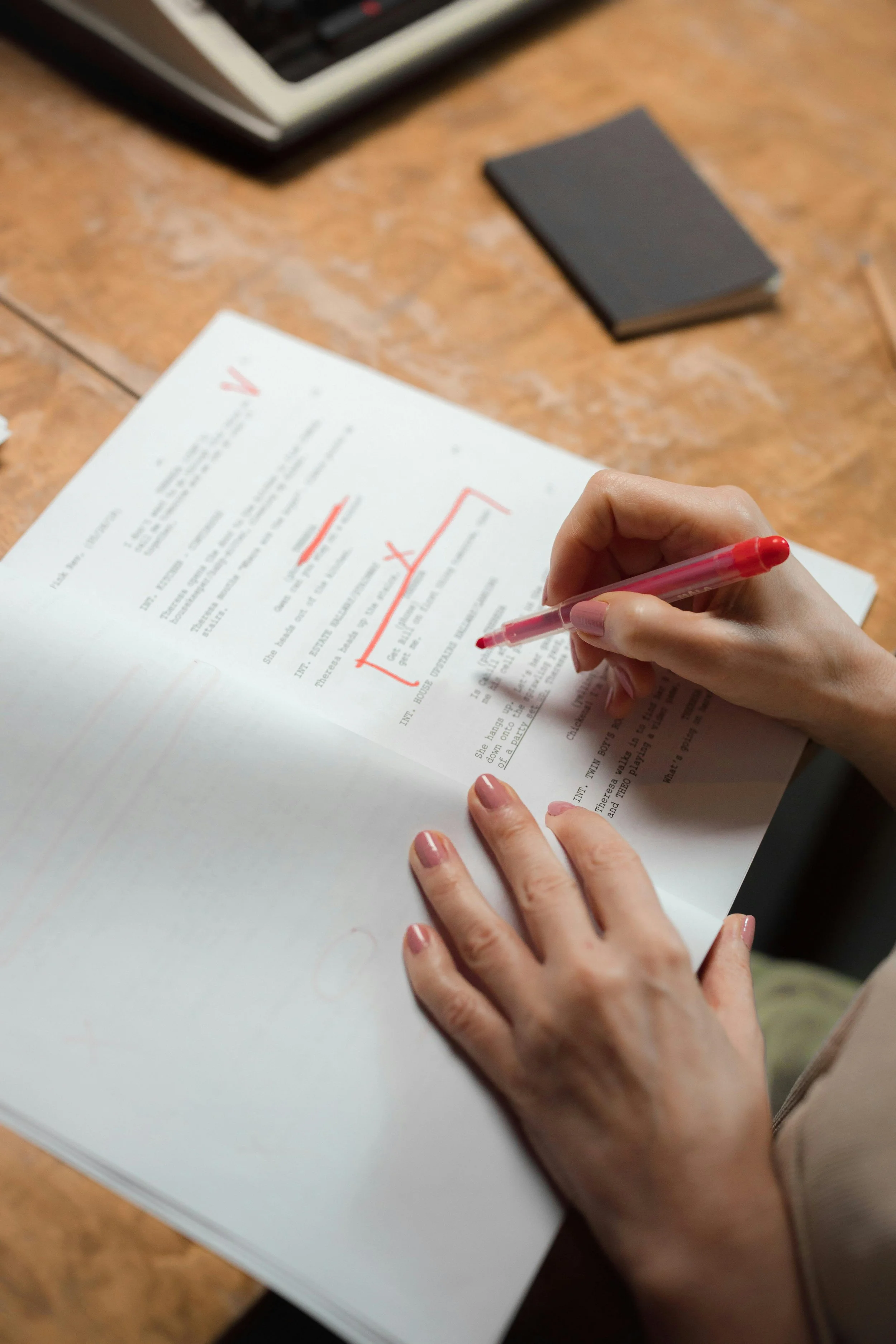 A person holding a red pen and marking a document on a wooden table. There are black and white books and a keyboard in the background.