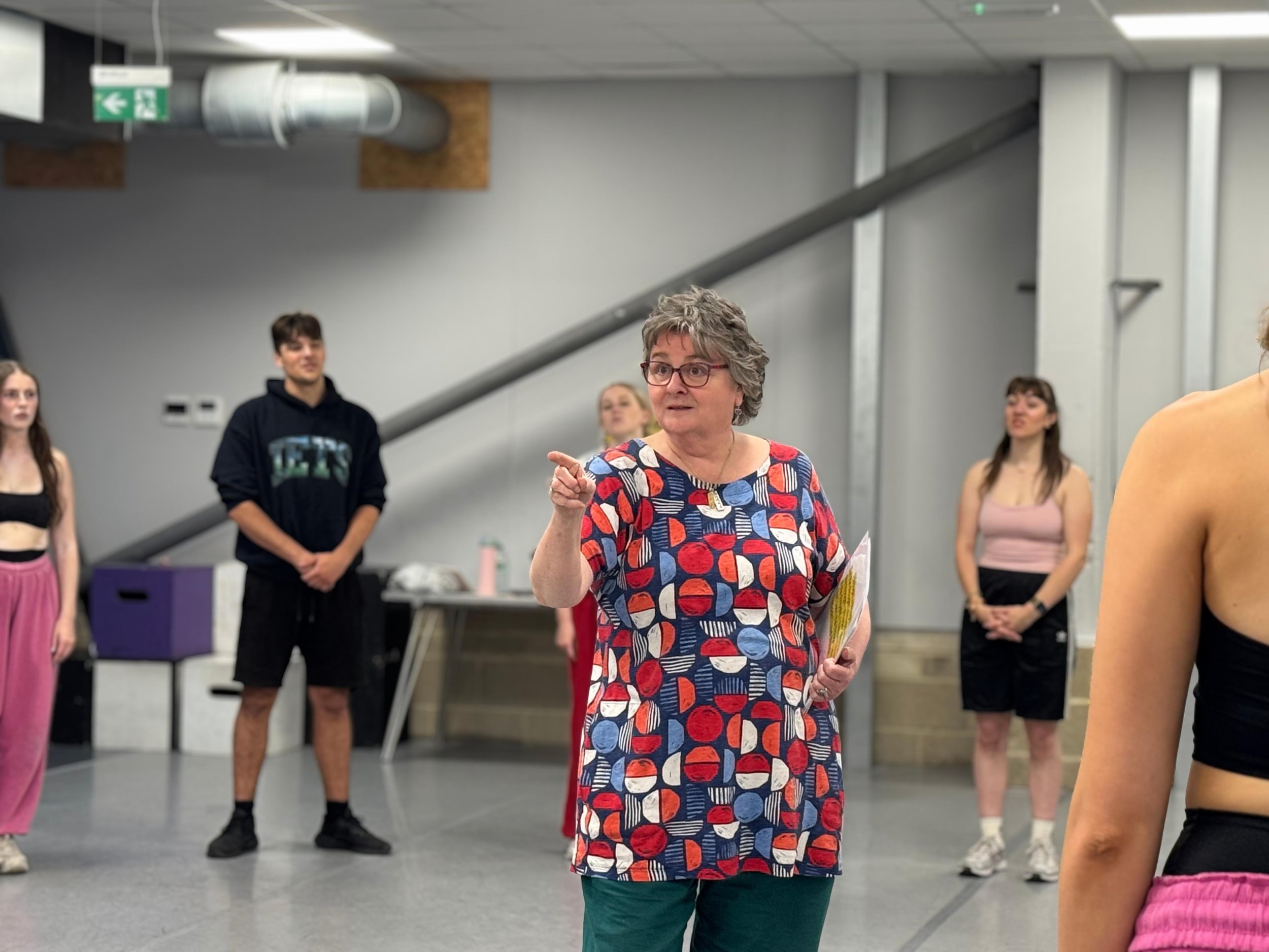 Sara Ford A woman with gray hair and glasses wearing a colorful patterned dress is pointing while speaking to a group in an indoor setting.