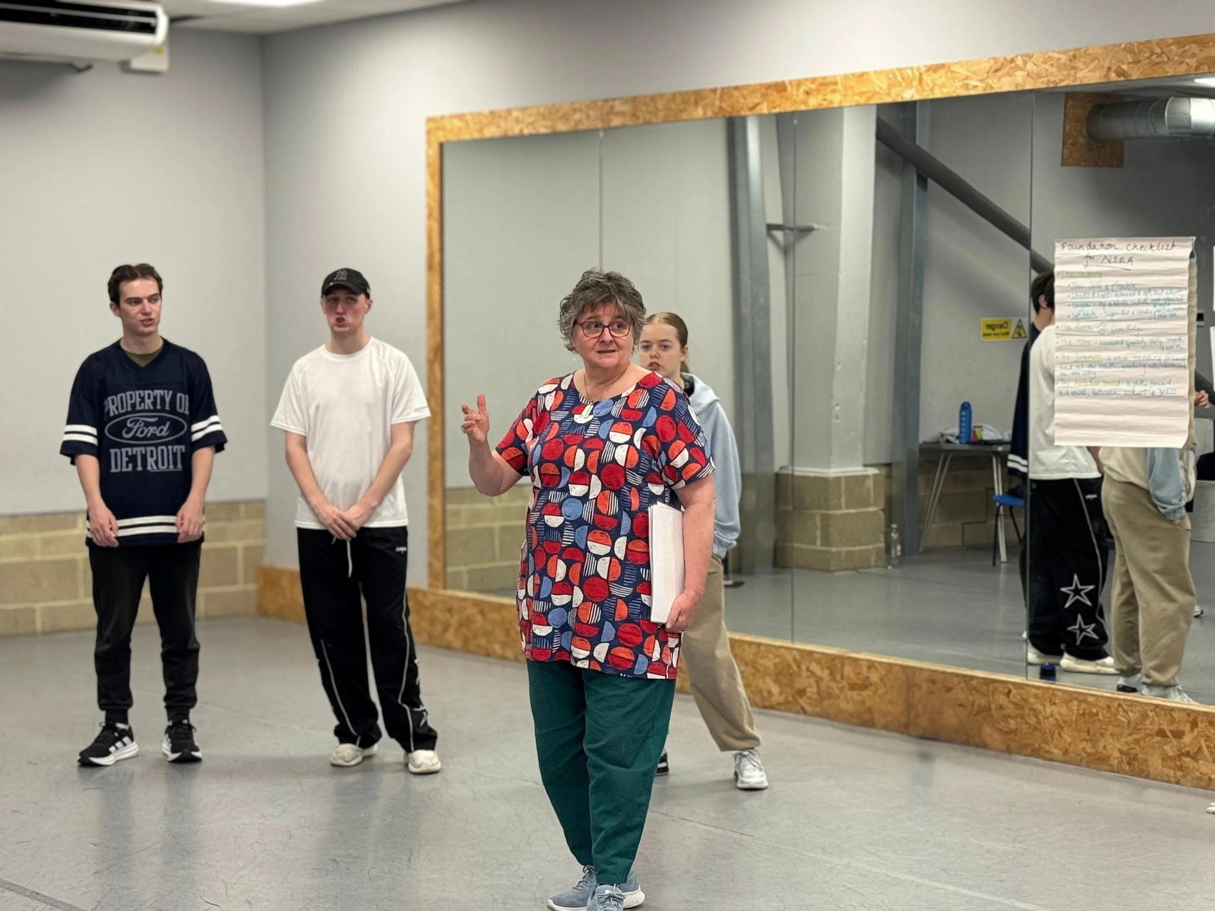 Older woman in glasses and colorful dress speaking to a group of young people in a room with a large mirror and a sheet of paper on an easel.