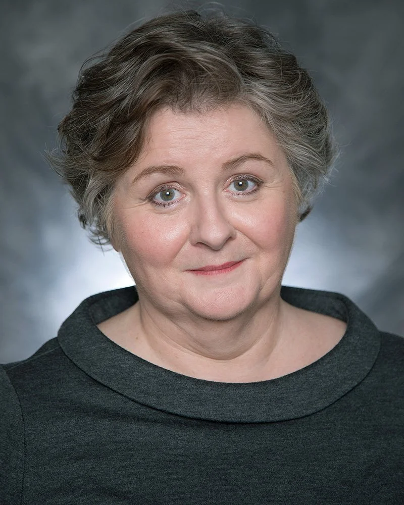 Sarah Ford with short gray hair and blue eyes, wearing a black top, looking directly at the camera against a blurred gray background.