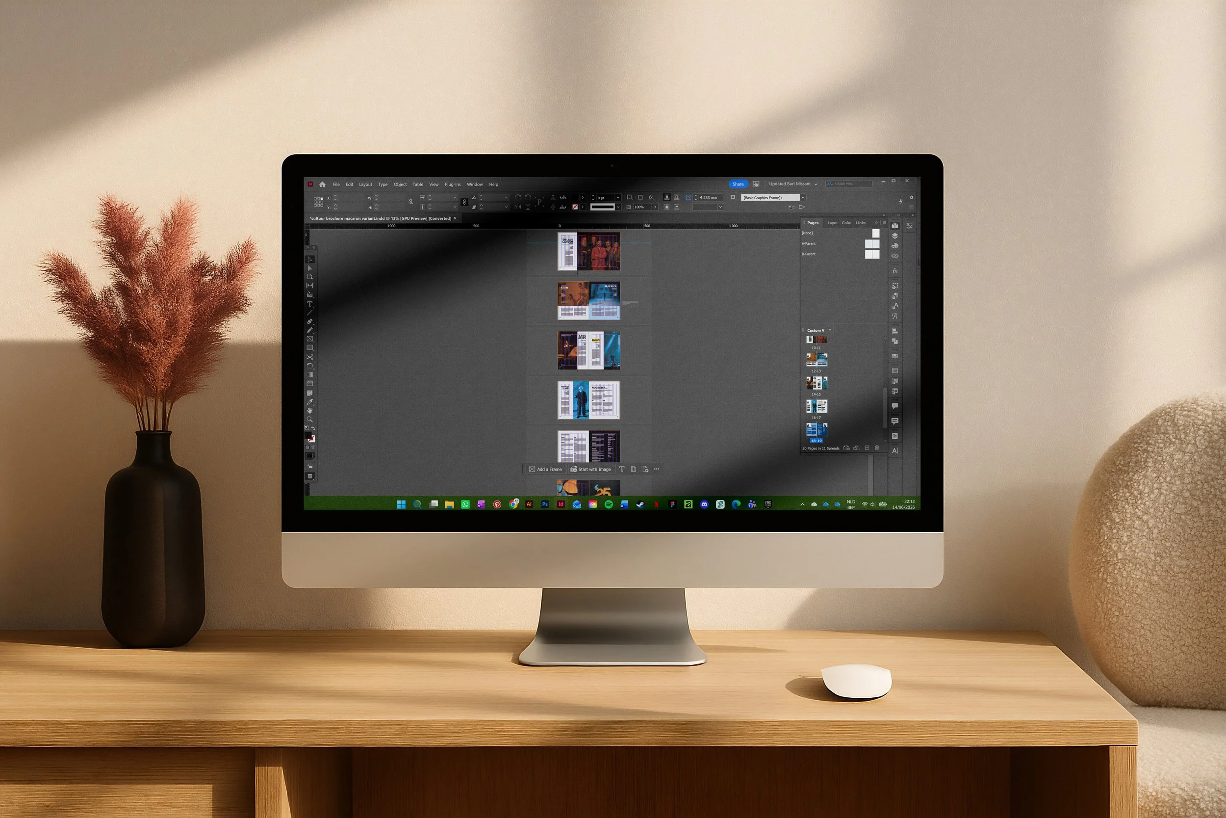 Een beeld van een iMac-computer op een houten bureau met een ontwerpsoftware op het scherm, naast een zwarte vaas met roze pampasgras en een beige tapijt op de grond.