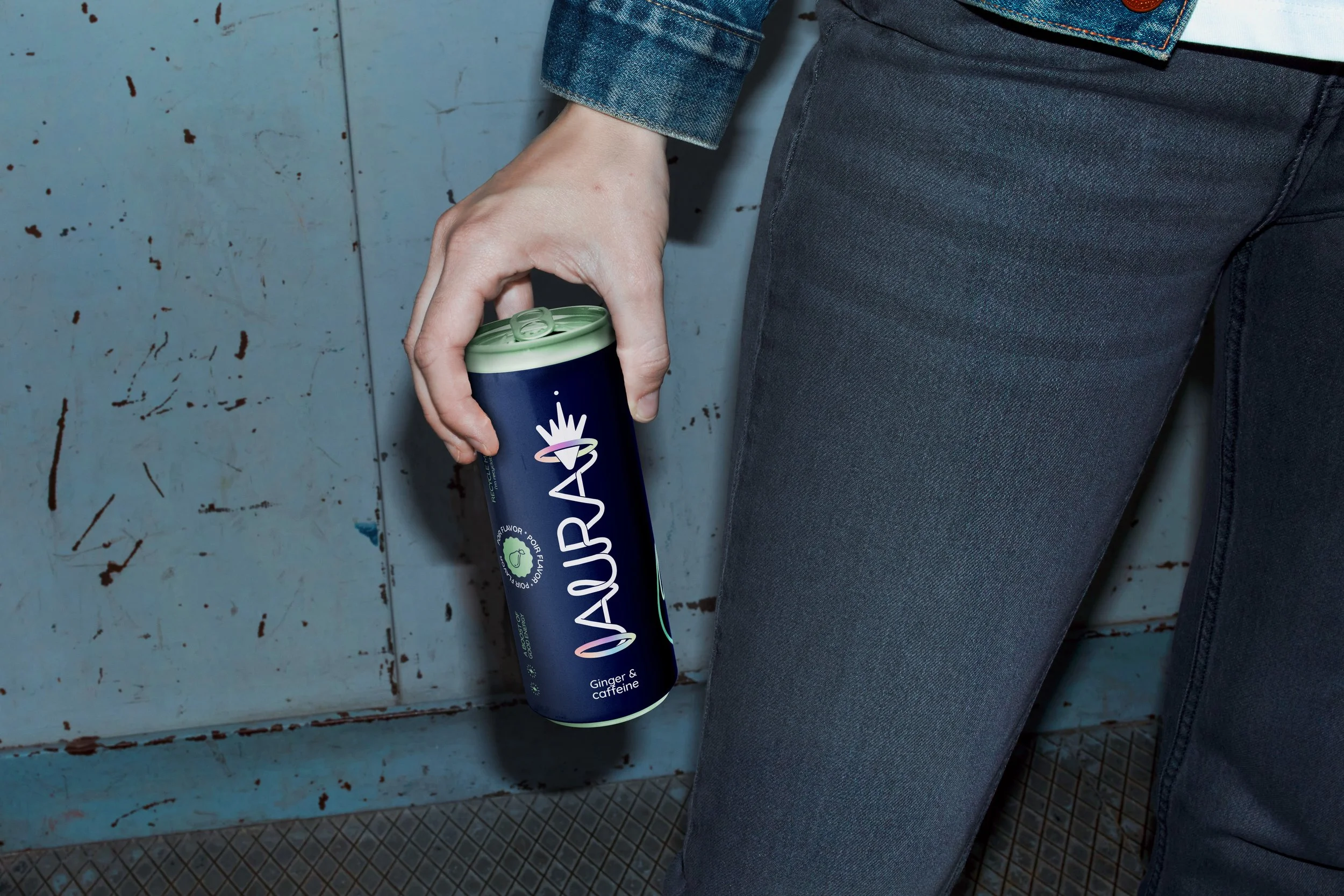 Persoon in blauwe spijkerjas en donkere broek die een blikje energiedrank vasthoudt, tegen een blauwe muur met roestplekken.
