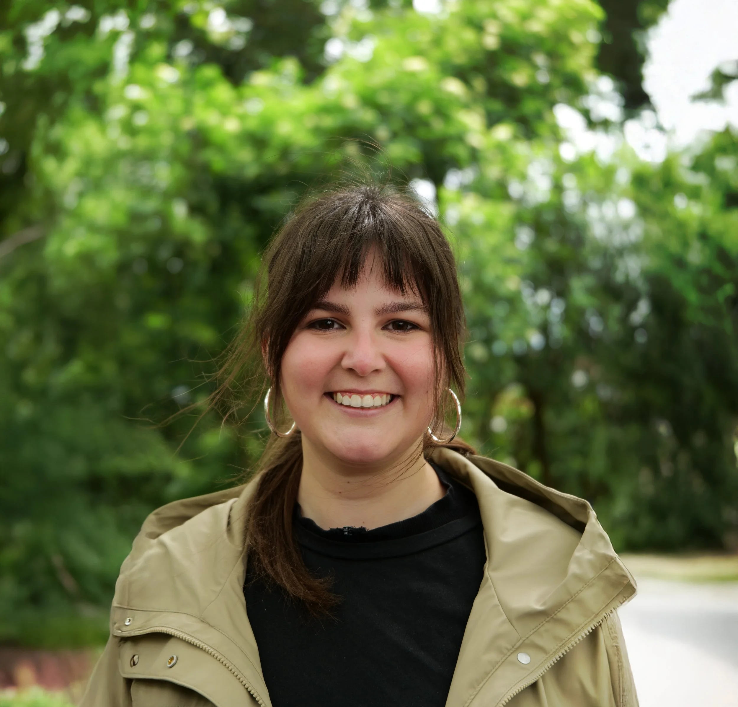 Close-up van een lachende vrouw met bruin haar, grote zilveren oorbellen, zwarte top en beige jas, buiten met groene bladeren op de achtergrond.