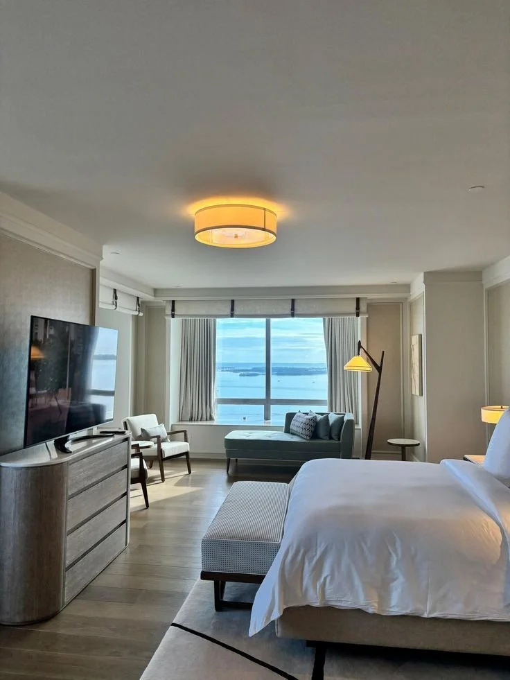 A hotel room with a large window overlooking a body of water. The room features a flat-screen TV, a bed with white linens, a small seating area with a blue sofa, a floor lamp, and a beige ottoman at the foot of the bed.