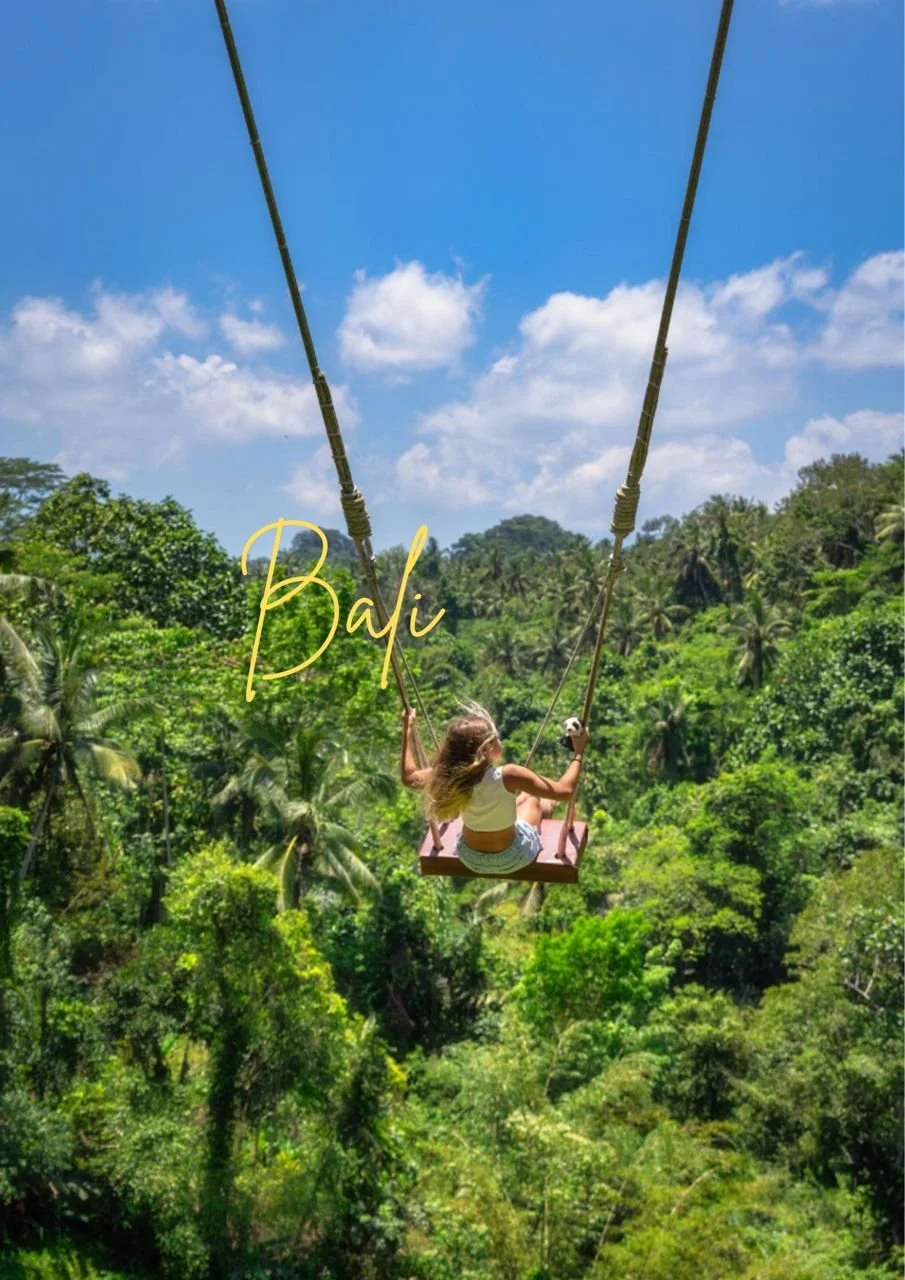 A woman on a swing hanging high above a lush green tropical forest under a bright blue sky with scattered clouds in Bali.