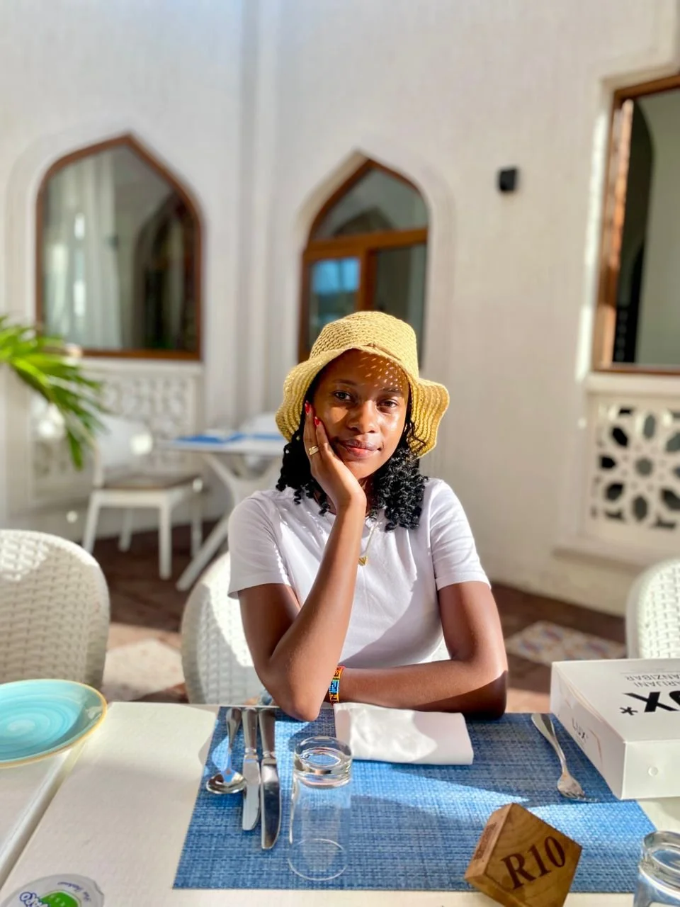 A woman sitting at a table with a blue tablecloth, wearing a straw hat and white shirt, in a well-lit room with arched windows.