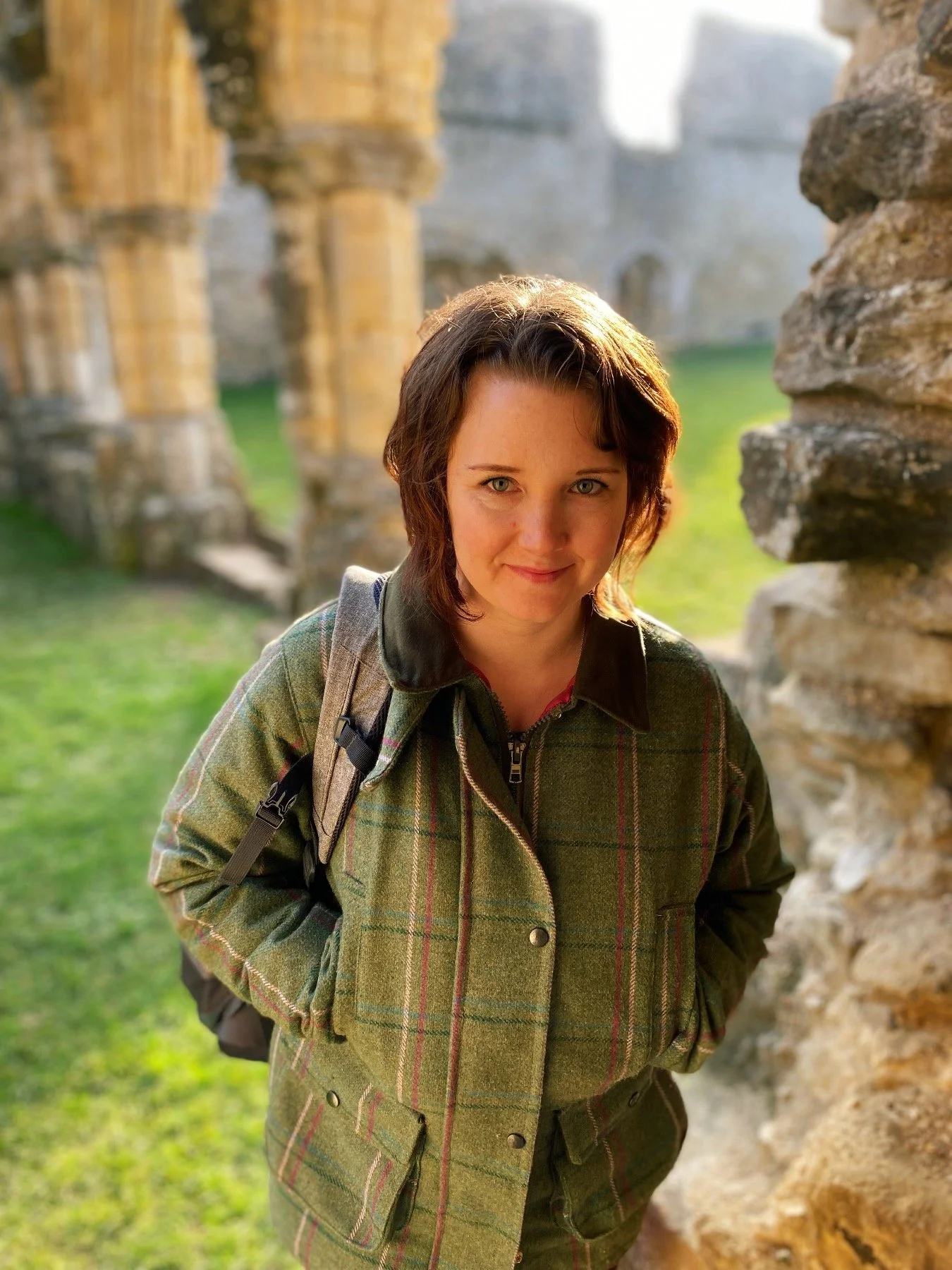 A woman with short brown hair and blue eyes wearing a green plaid jacket and carrying a backpack, standing outdoors near stone ruins with sunlight illuminating her face.