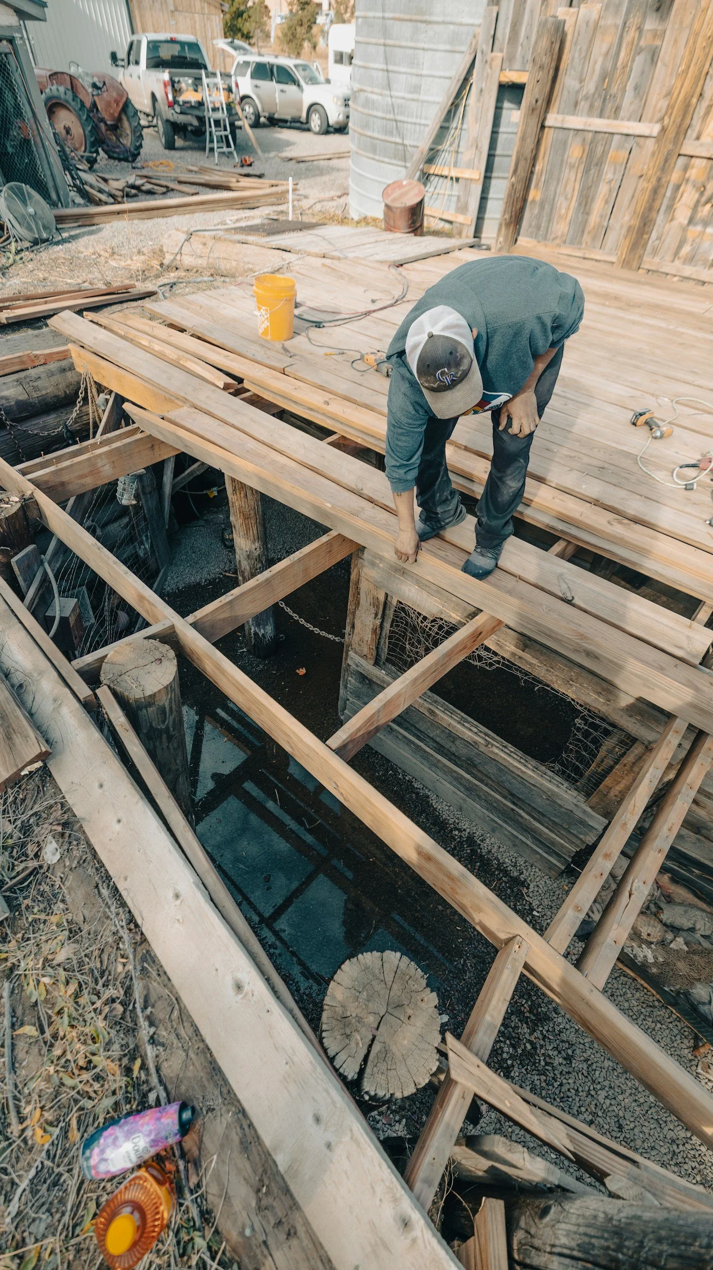 A man on a construction site working on a wooden deck or platform, with building materials, tools, and parked vehicles in the background.