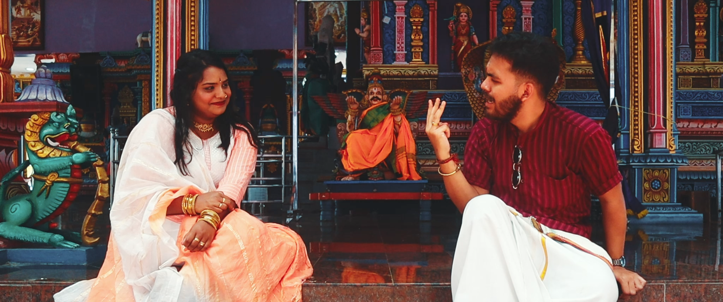 A woman and man sitting on the floor inside a Hindu temple, having a conversation. The woman is dressed in traditional white and orange attire with gold jewelry, while the man is wearing a red shirt and white dhoti. The temple interior has colorful deity statues and intricate decorations.