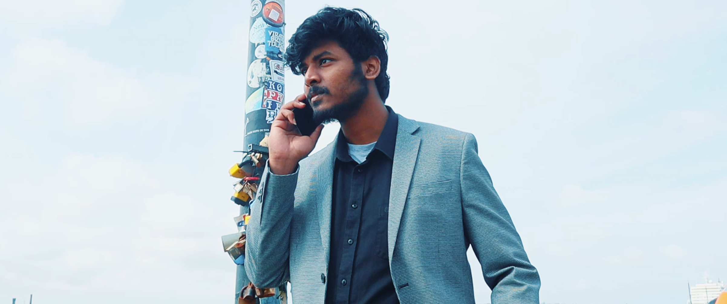 A young man with curly hair and a beard in a gray blazer and black shirt talking on a cell phone outdoors, standing next to a utility pole covered with stickers, against a cloudy sky.
