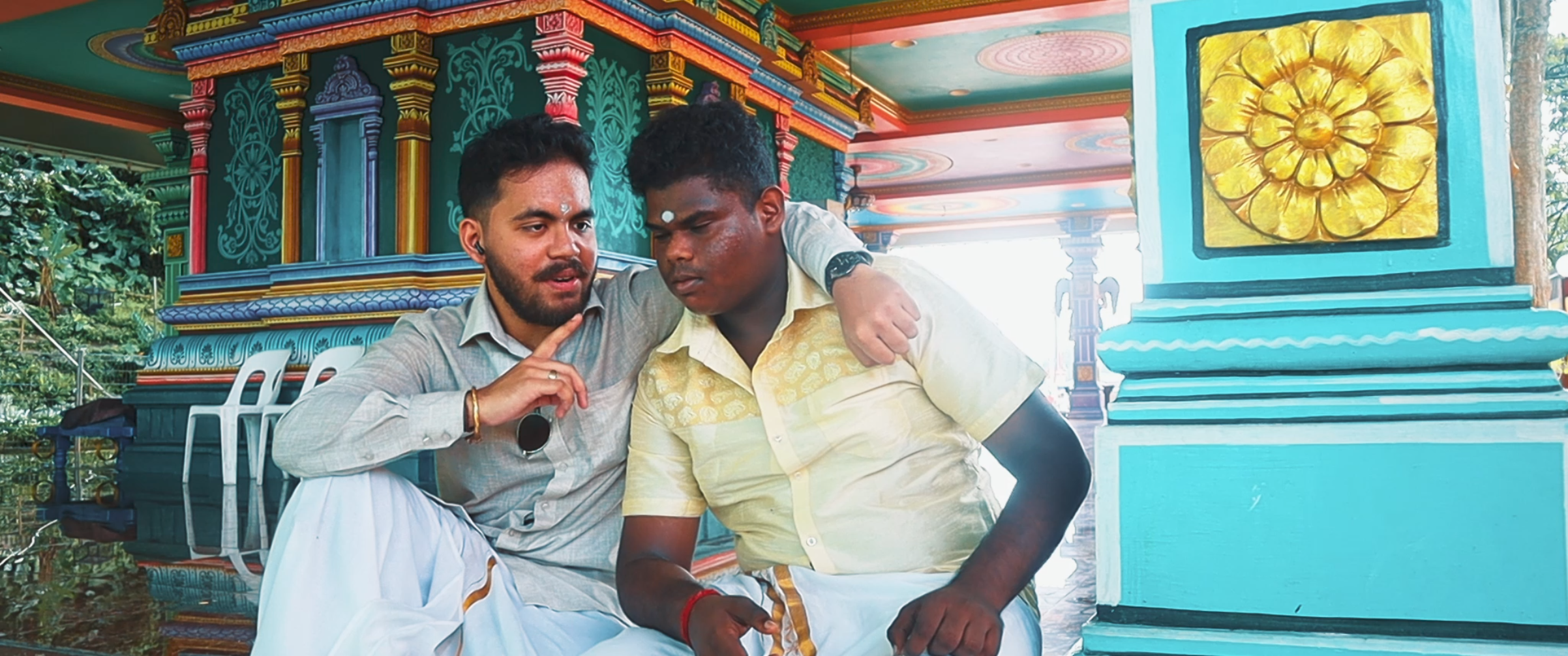 Two men sitting closely together, one with a hand on the other's shoulder, inside a colorful temple with ornate decorations and a yellow floral artwork on the wall.