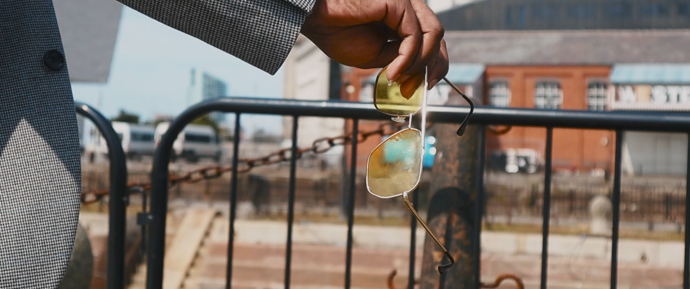 A person holding sunglasses outdoors with a cityscape in the background.