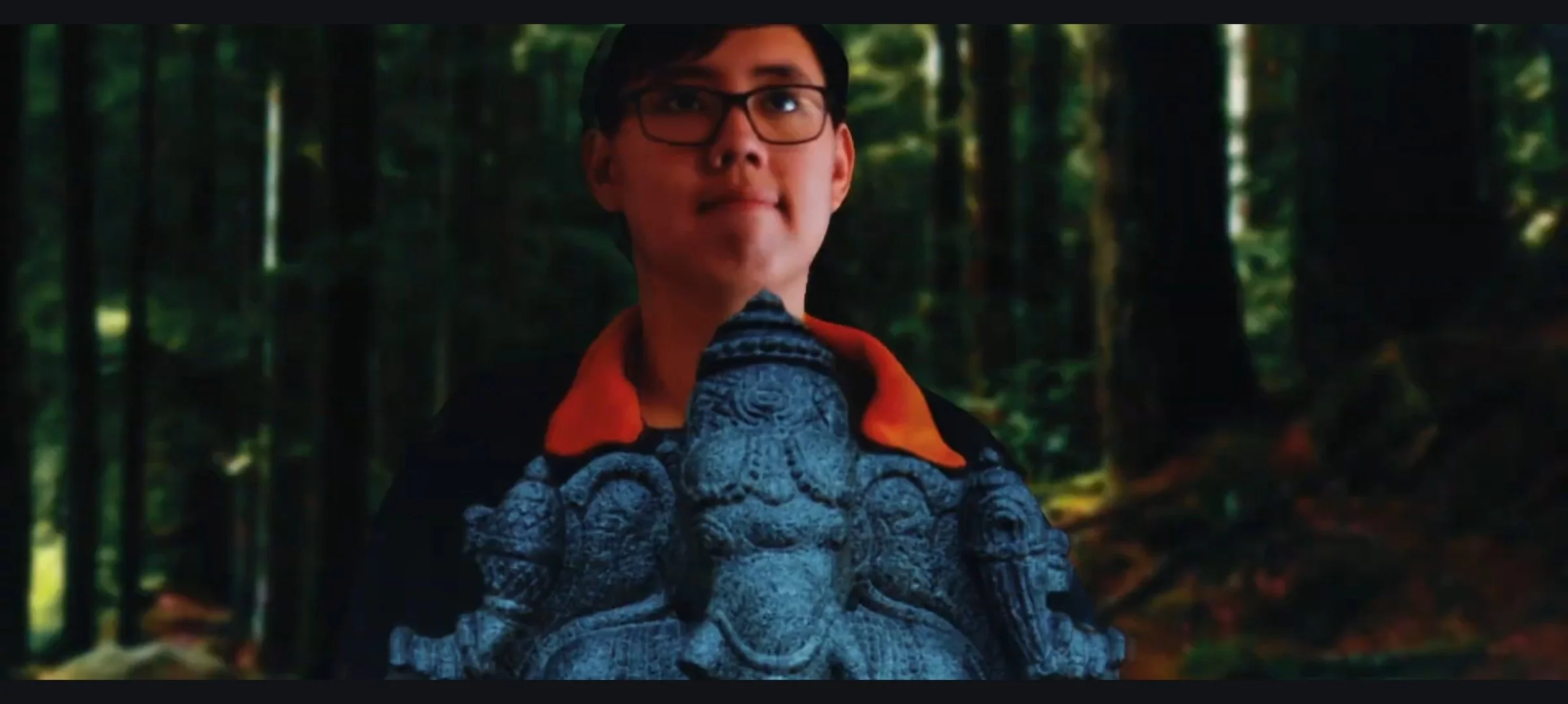 A young man with glasses and dark hair holding a carved stone sculpture of Lord Ganesha in a forest setting.