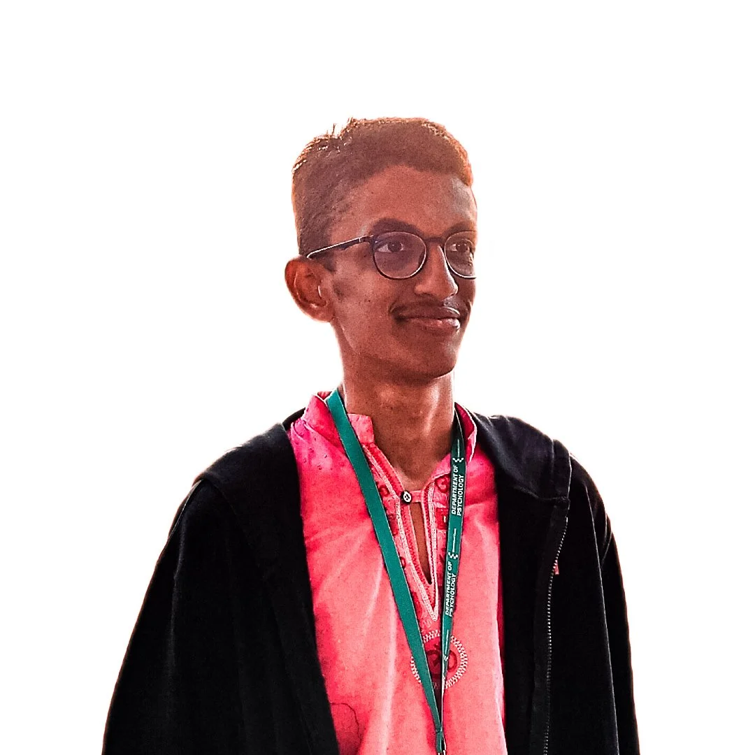 A woman with short hair wearing glasses, a pink shirt, and a black jacket, with a green lanyard around her neck, standing against a plain light background.
