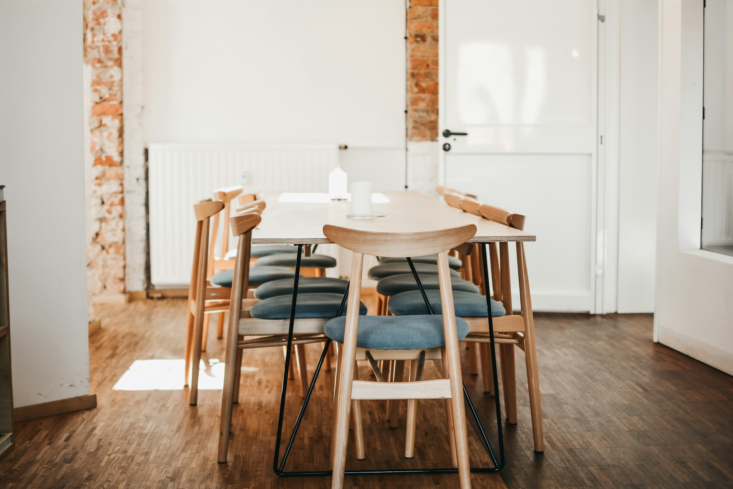 A dining room with a long wooden table surrounded by wooden chairs with gray cushions, modern decor, exposed brick accents, white walls, and hardwood flooring.