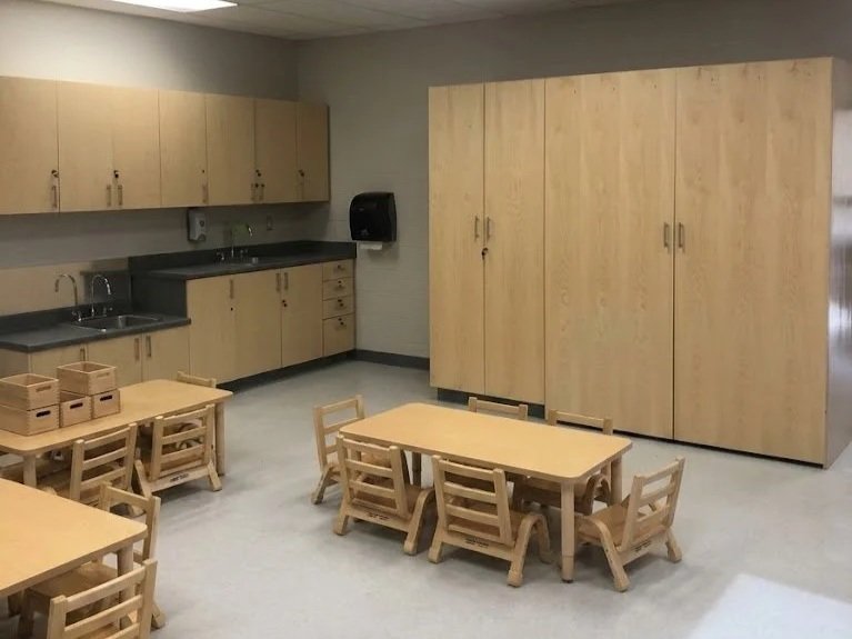 A classroom kitchenette with wooden cabinets, small tables, chairs, and storage bins.