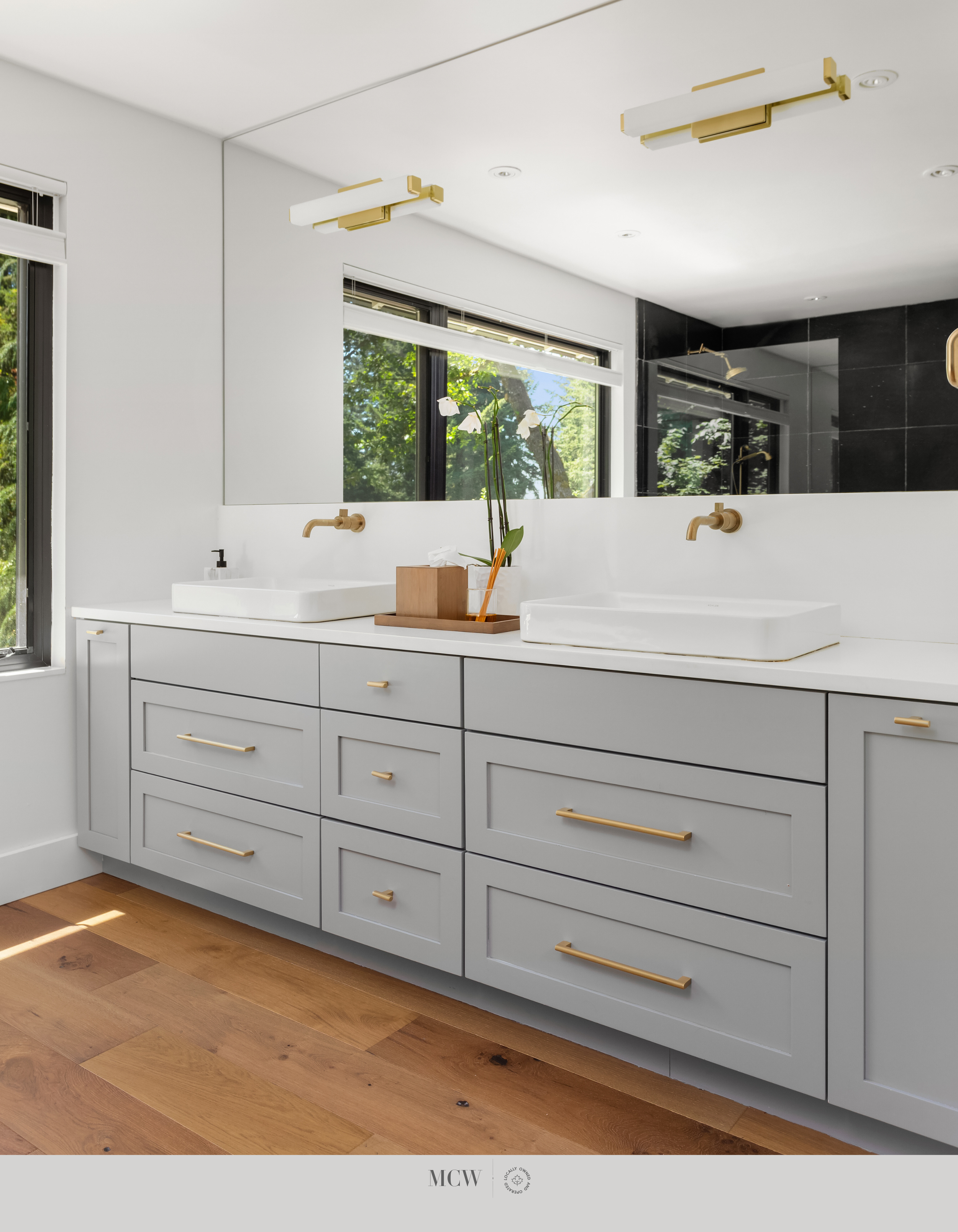 Modern bathroom with gray cabinetry, gold handles, white sinks, a wooden countertop with a flower arrangement, large window with trees outside, and a black-tiled shower area.