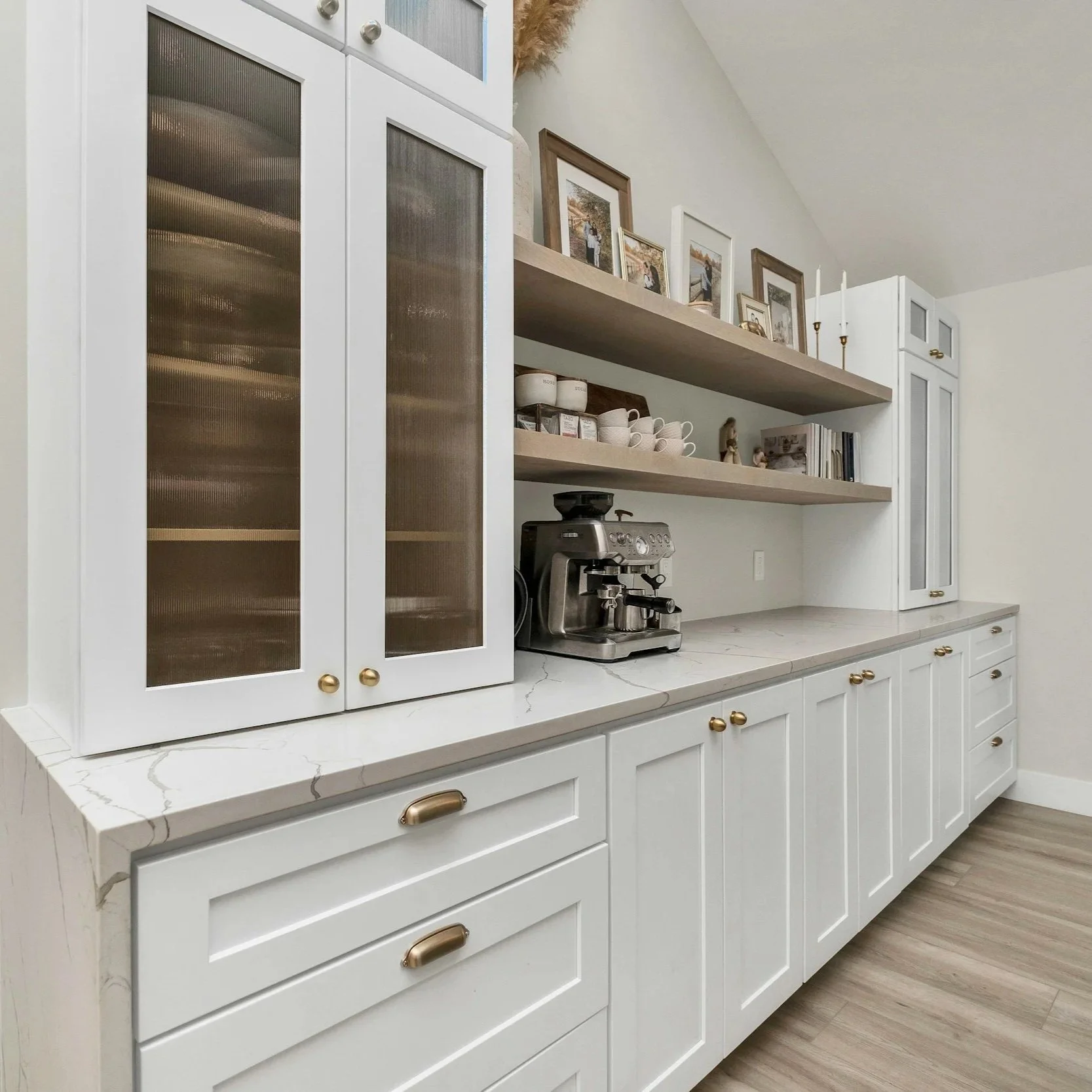 Kitchen countertop with white cabinetry, wooden open shelves with framed family photos, cups, and a small decorative figurine. A coffee machine is on the counter. The kitchen has a light color scheme with a wood floor.