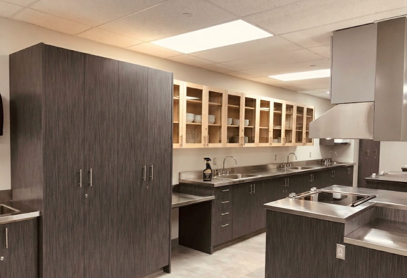 Commercial kitchen with gray cabinets, open wooden shelves with dishes, and a stainless steel countertop with sinks