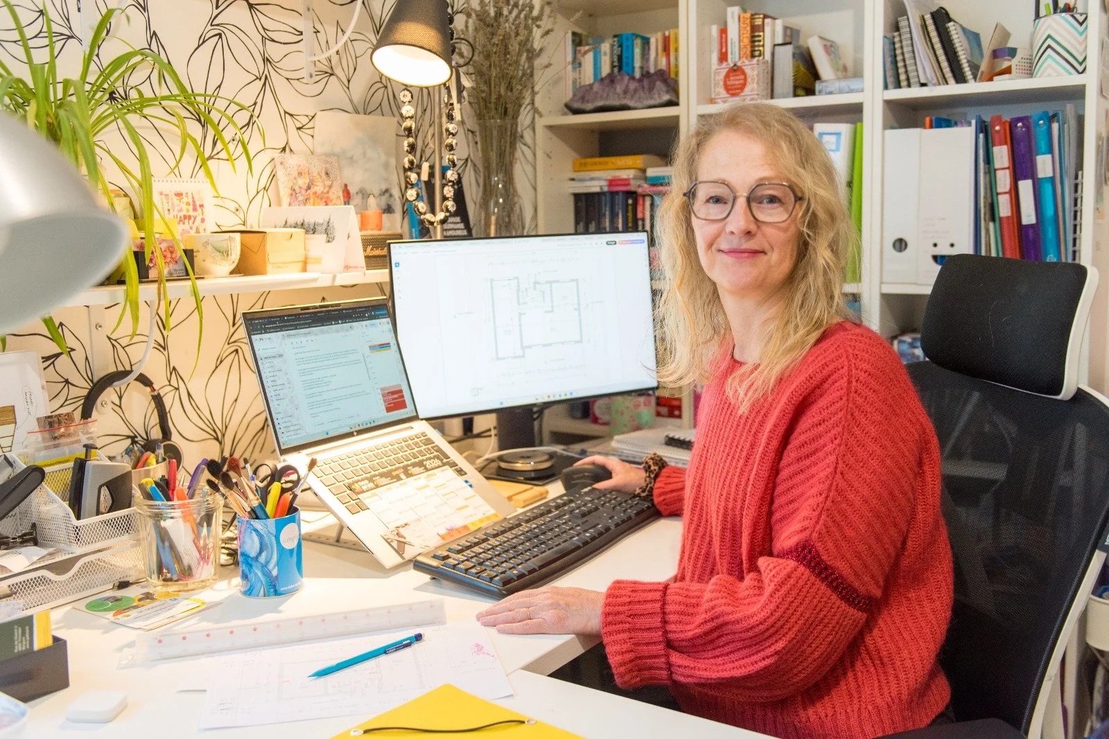 Une femme assise à un bureau avec deux écrans d'ordinateur, entourée de fournitures de bureau et de livres, dans un espace de travail organisé et décoré.
