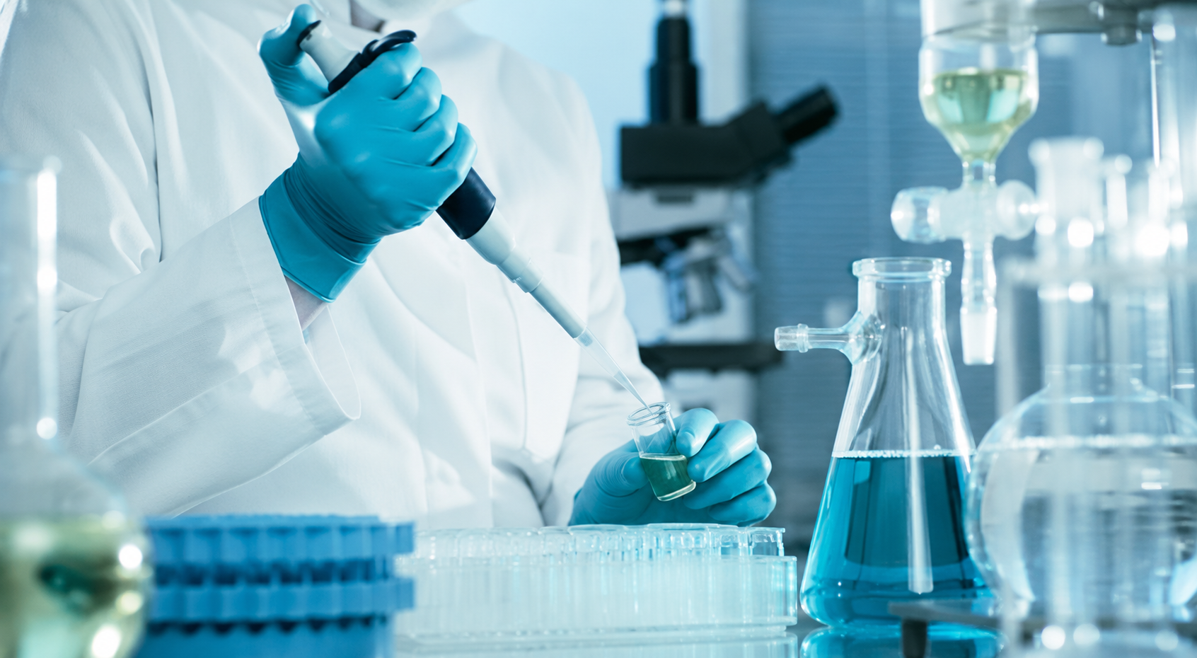 A scientist wearing blue gloves and a white lab coat using a pipette to transfer liquid into a test tube in a laboratory with glassware and equipment.