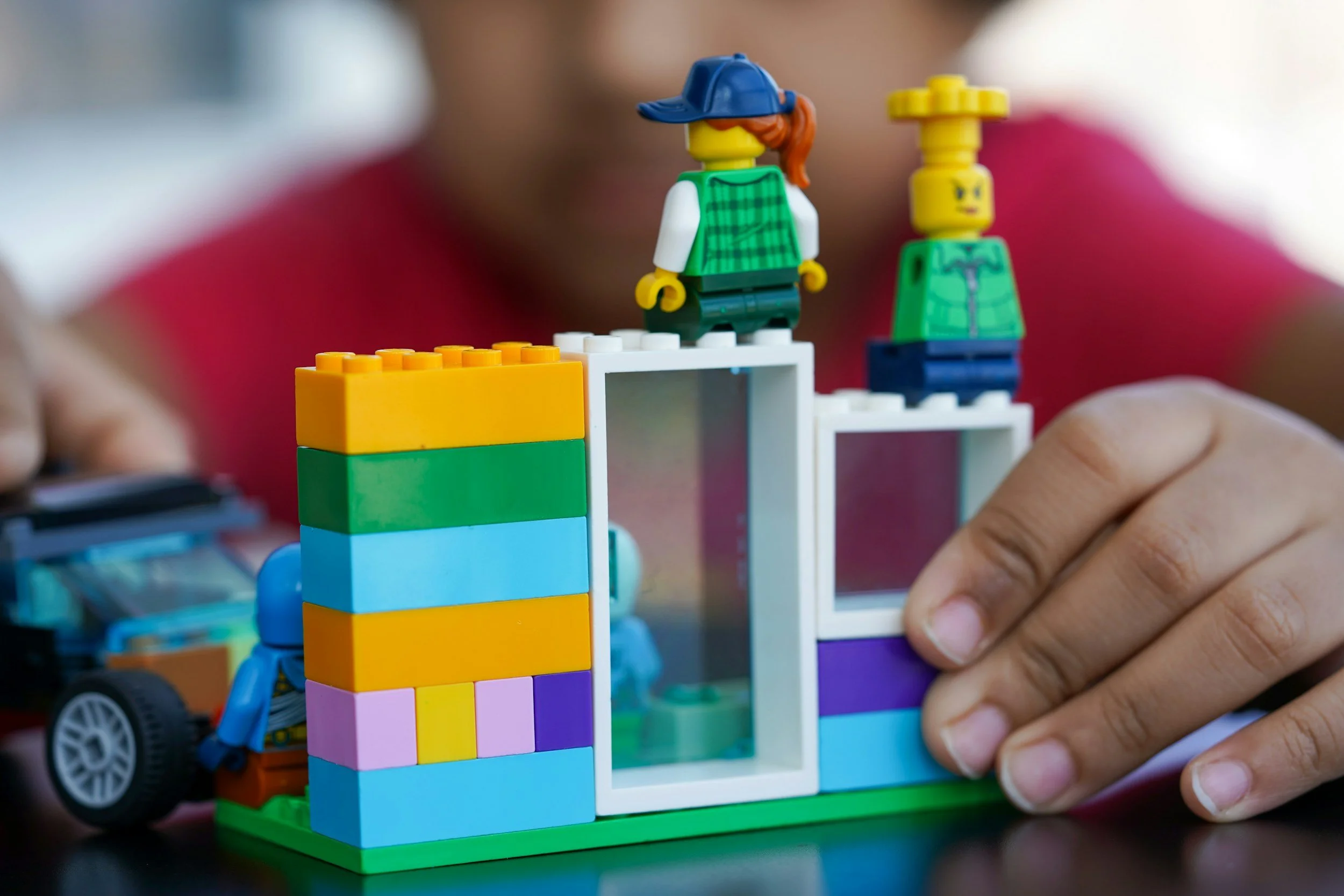 Close-up of LEGO figures and colorful building blocks arranged on a table, with a person's hand adjusting a piece.