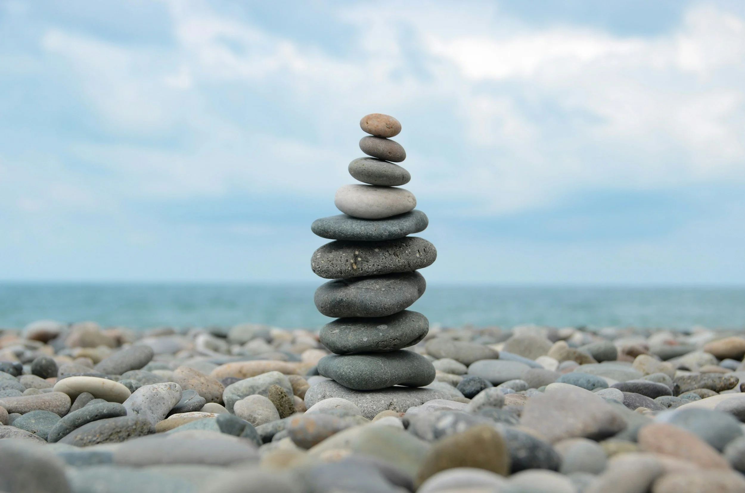 Stack of seven smooth stones balanced on top of each other on a pebble-covered beach with a cloudy sky in the background.